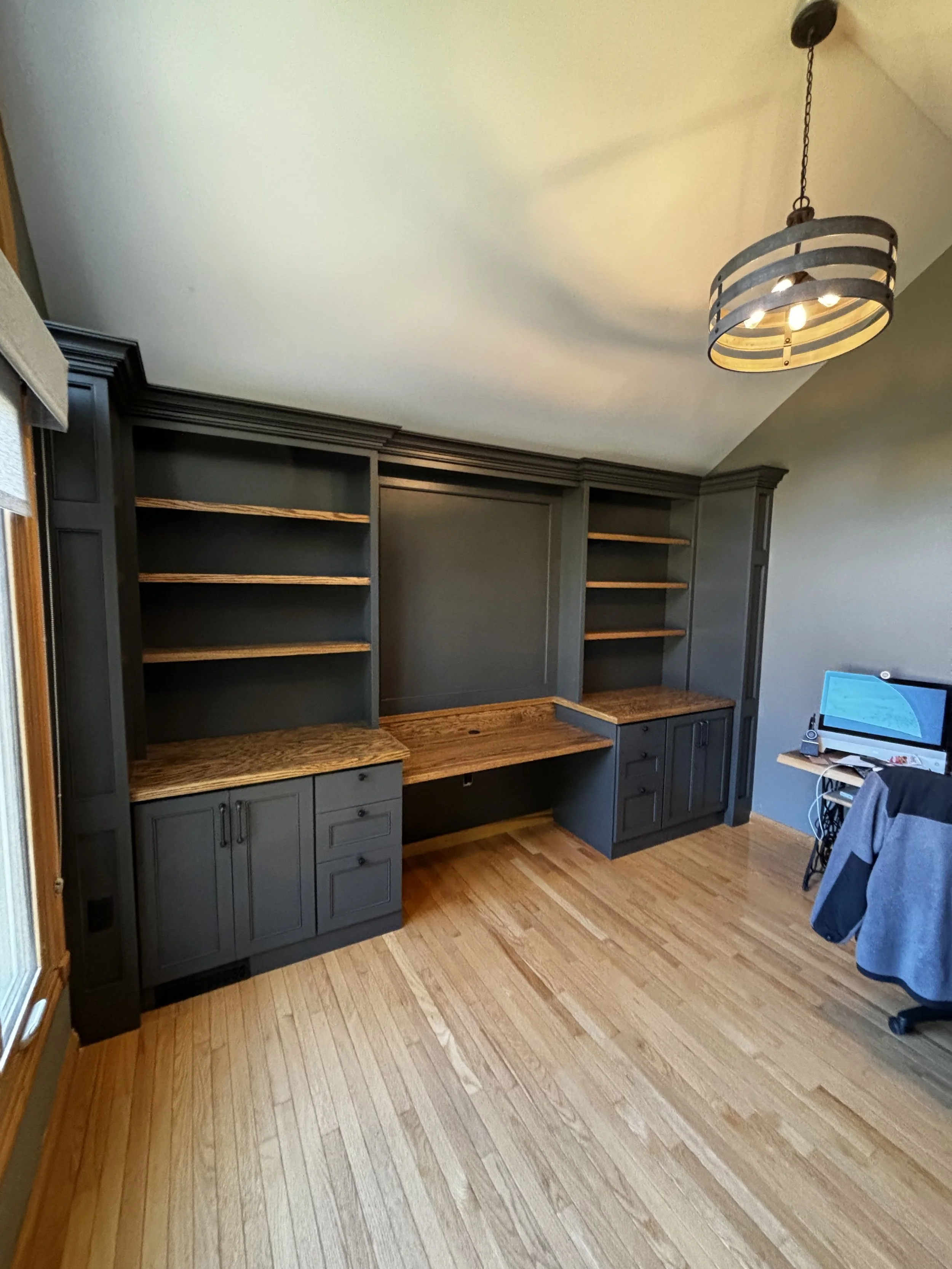 Room with wooden floor, dark gray built-in bookshelf and desk, ceiling light fixture, and a computer on a small table with a jacket hanging on a chair.