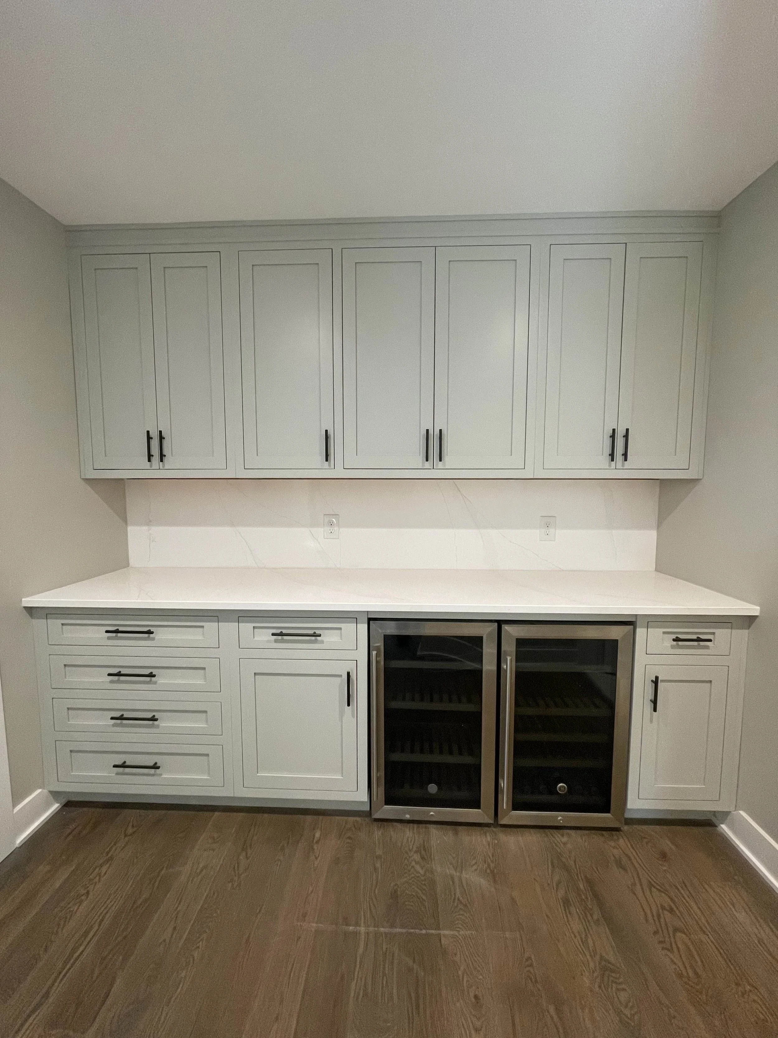 A kitchen cabinet with closed doors, a marble countertop, and a wine cooler beneath it. The cabinet is painted in a light color and has black handles. The wall behind the cabinet is painted light gray, and there are outlets and a marble backsplash vi