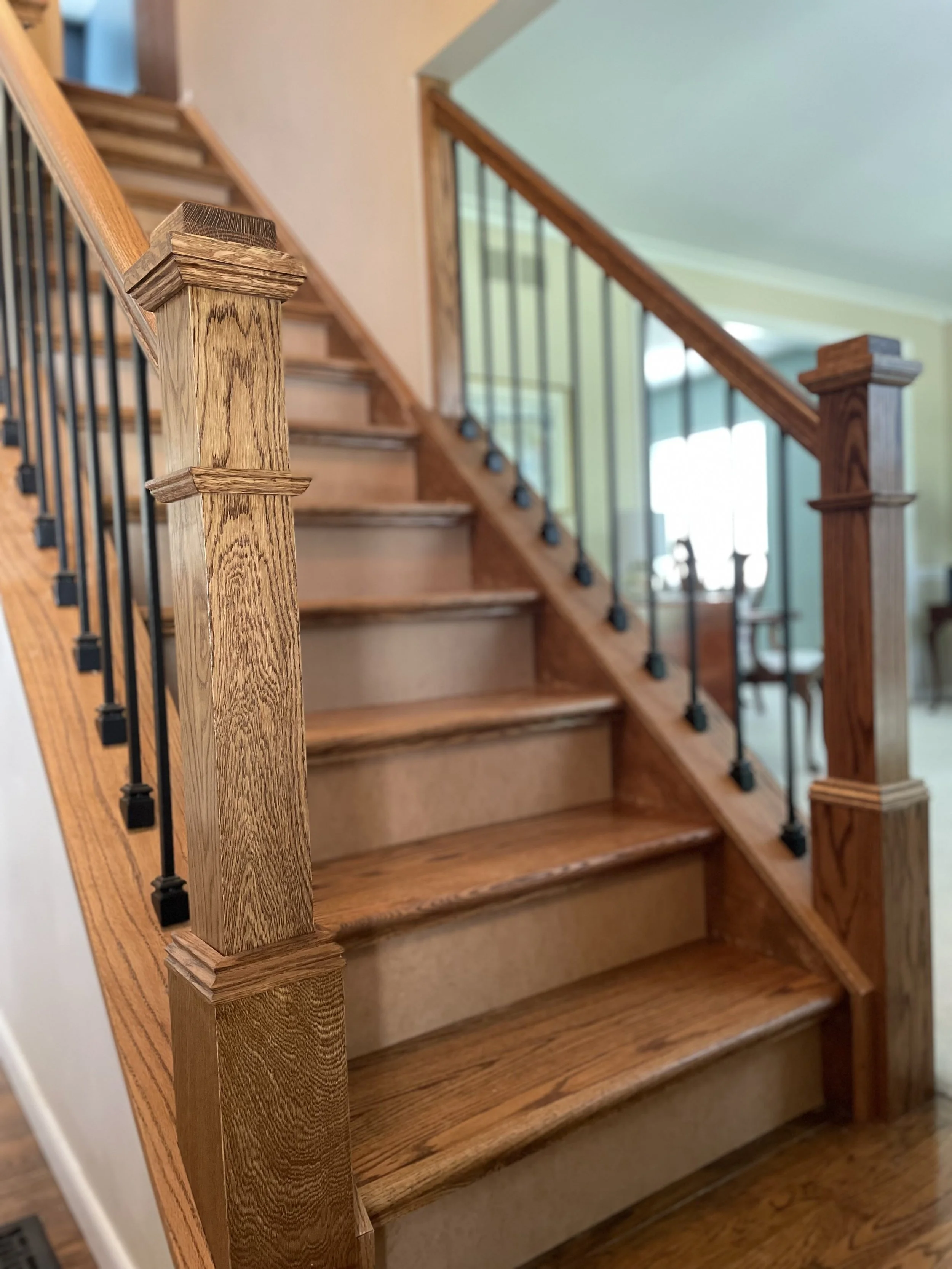 Close-up view of a wooden staircase's newel post and handrail with black metal balusters in a home interior.
