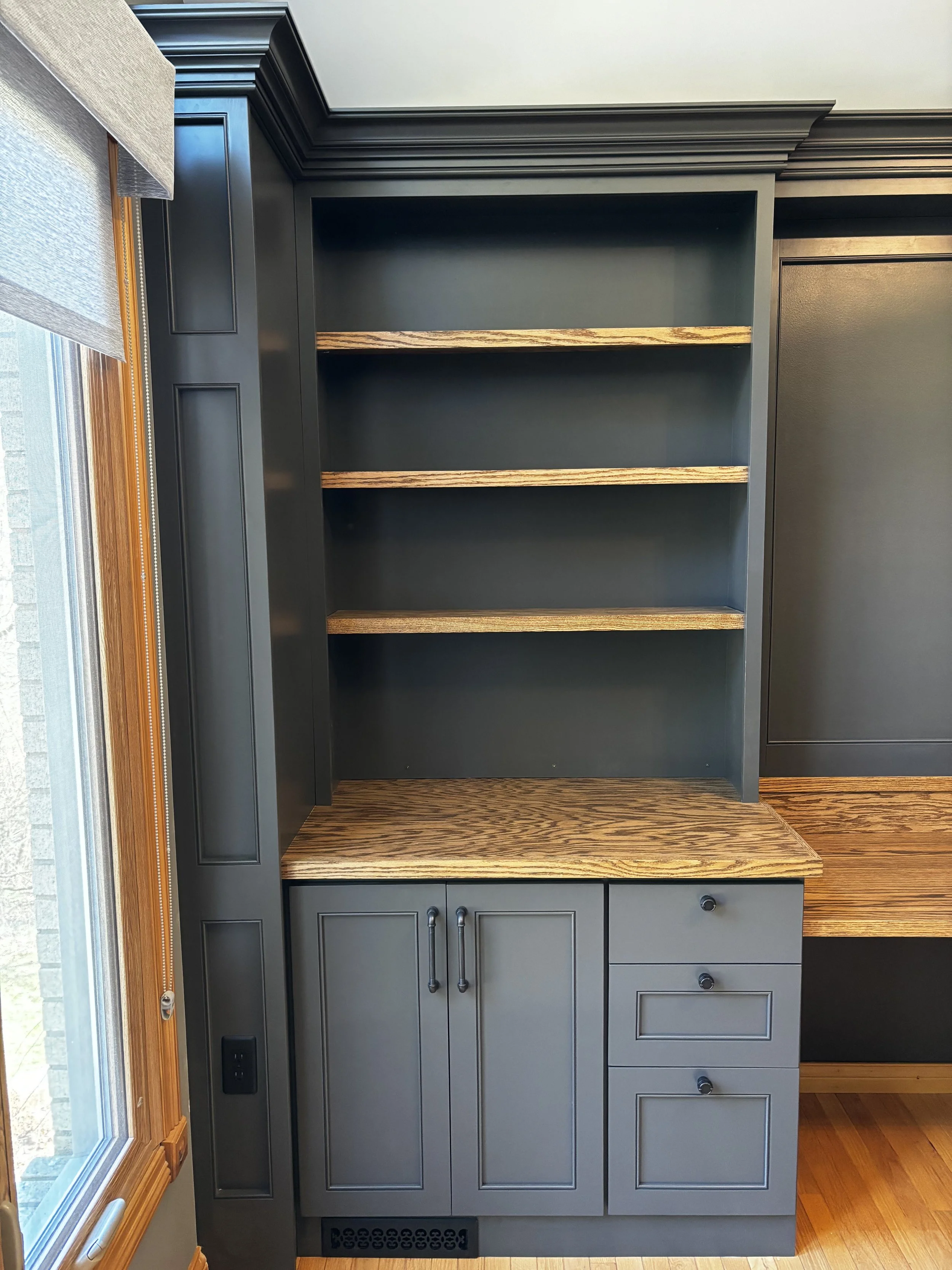 A built-in kitchen cabinet with dark gray finish, featuring three open shelves with wooden surfaces, a closed cabinet with double doors, and a small drawer with black knobs, positioned next to a window with wooden trim and beige blinds.