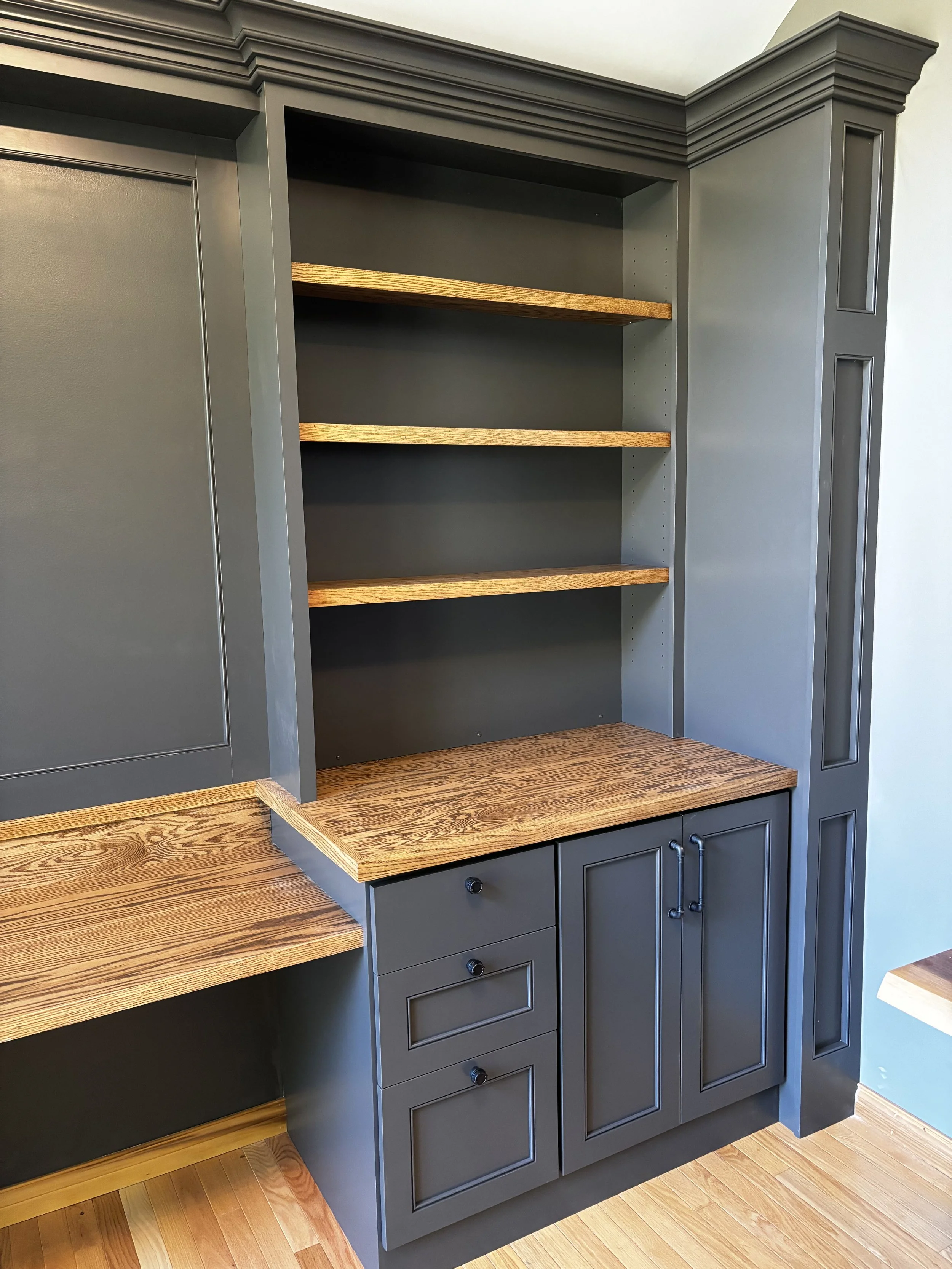 A kitchen cabinet with gray painted doors and trim, featuring open shelving with natural wood shelves, connected to a wooden countertop matching the floor.