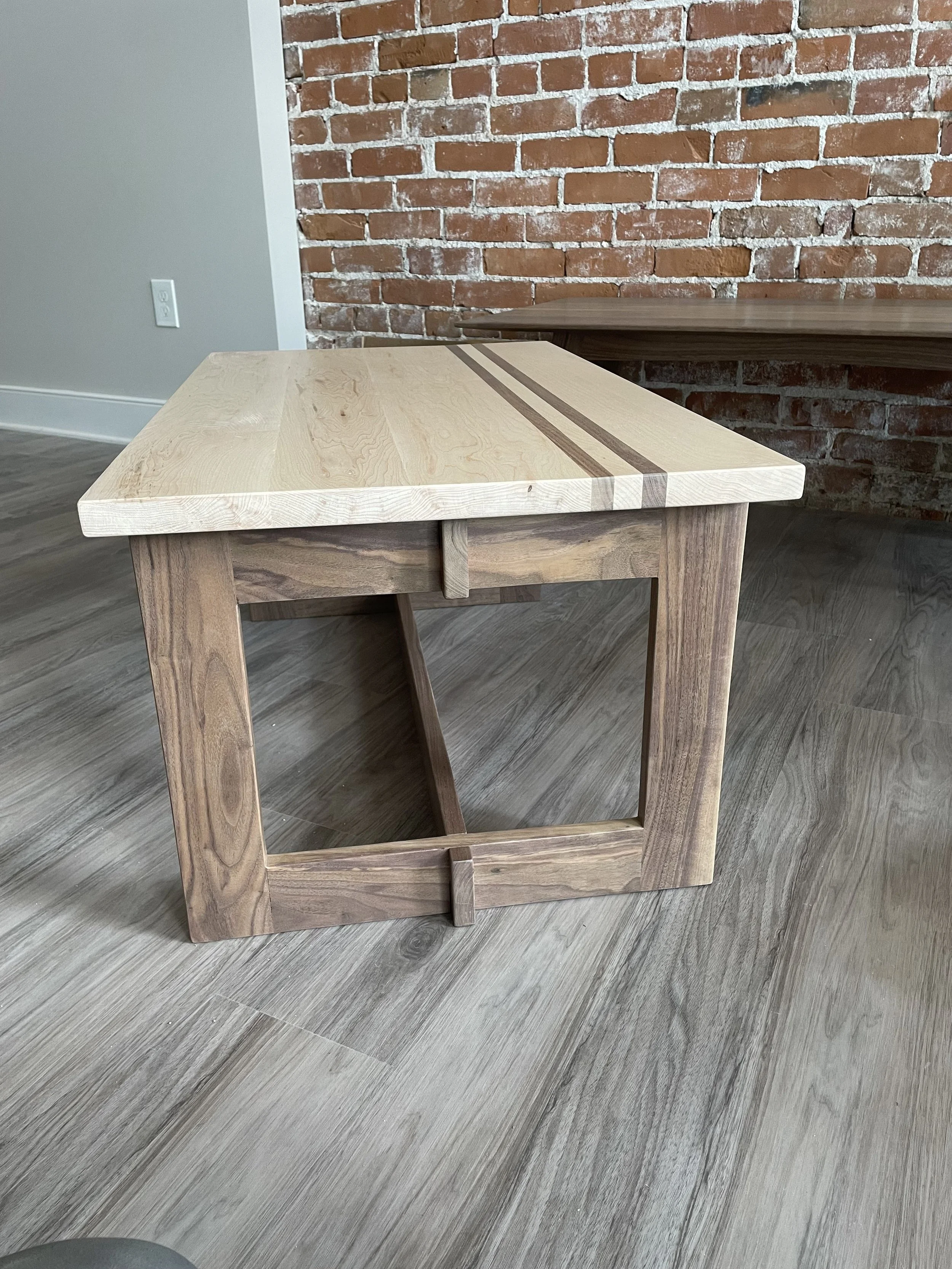 A wooden table with a light-colored top and darker striped inlay, placed on a gray wood-textured floor against an exposed brick wall background.