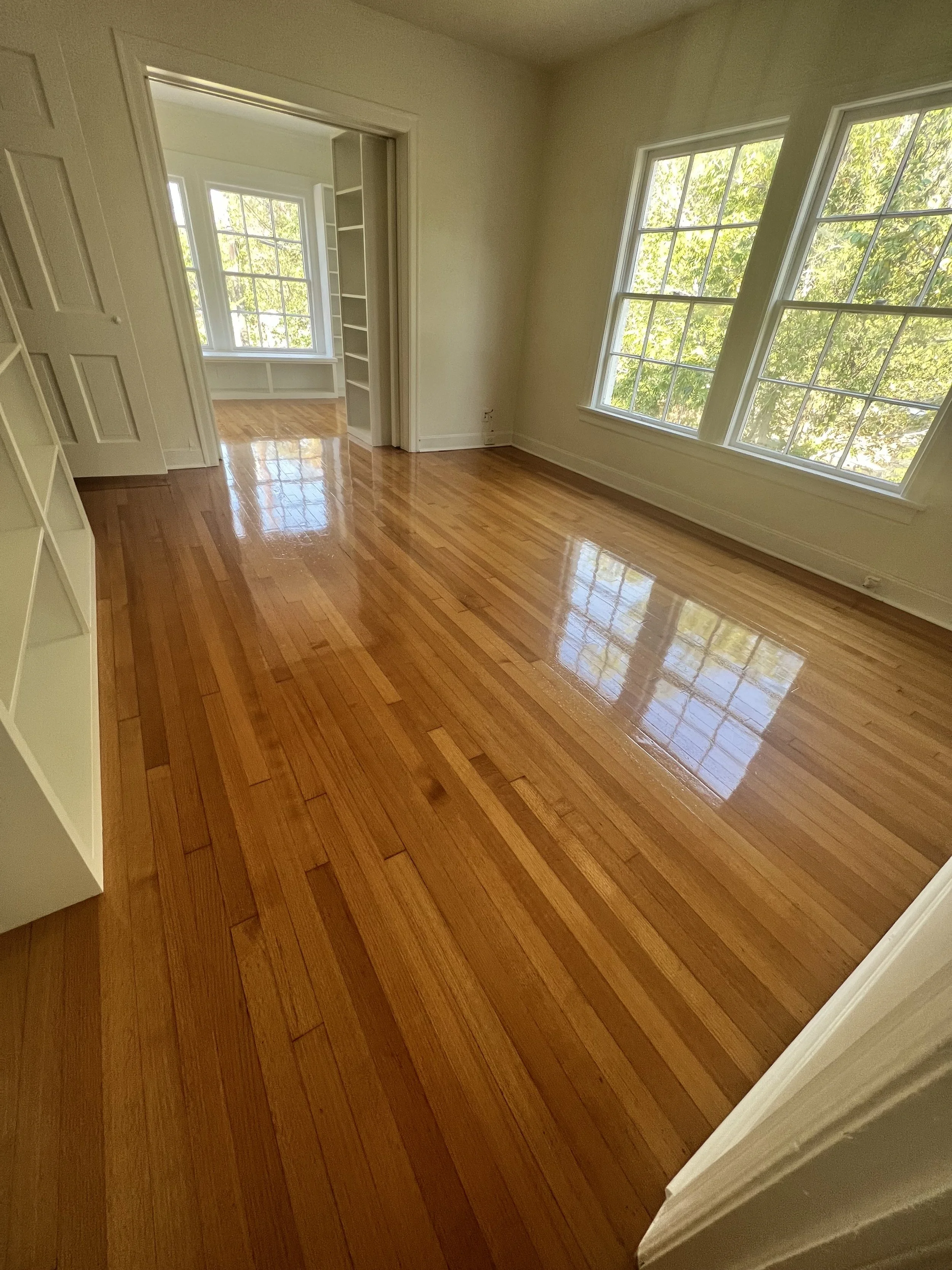 A bright empty room with polished hardwood floors, large windows letting in natural light, and a doorway leading to a sunlit room with more windows and built-in shelves.