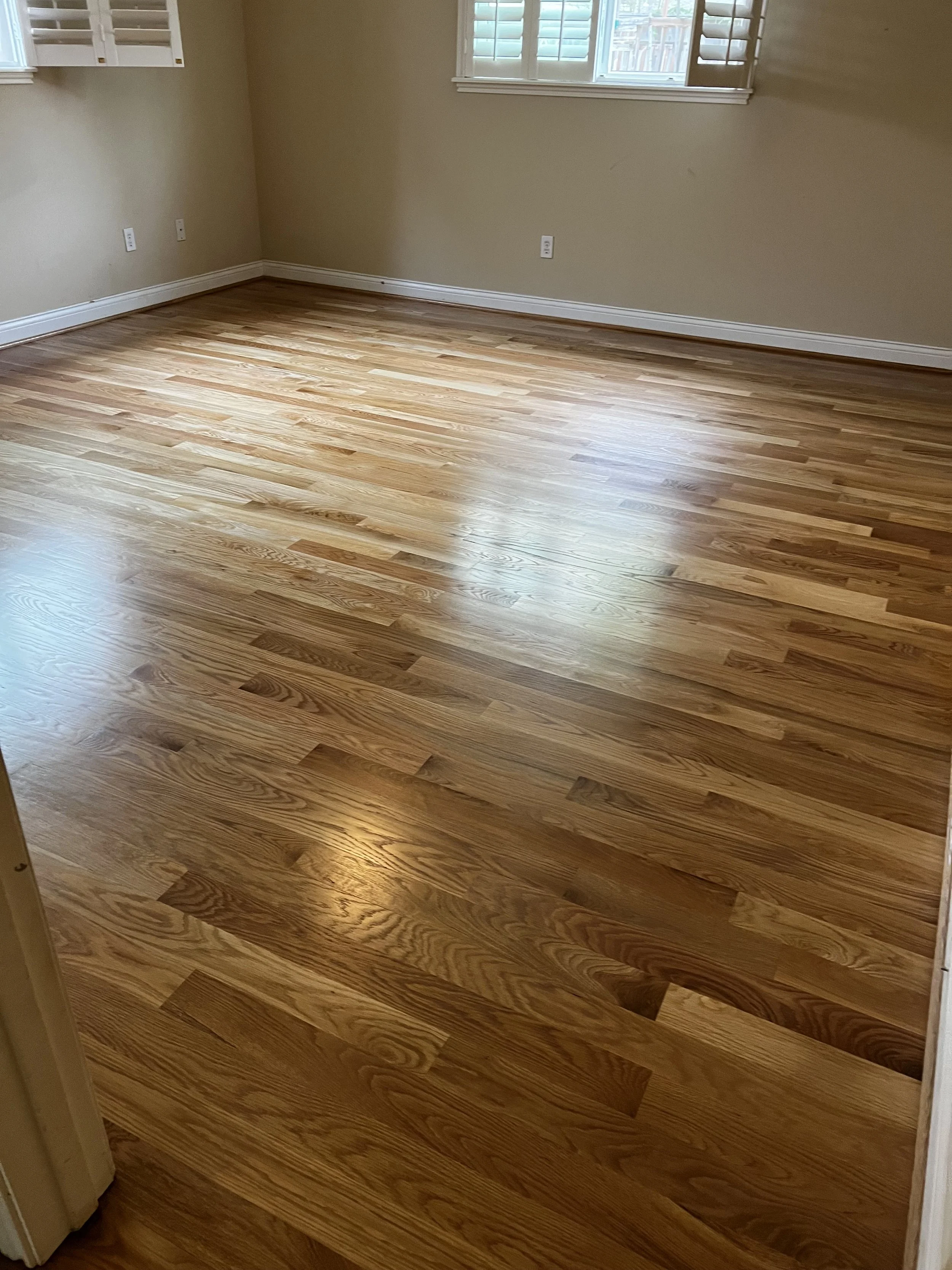 Empty room with hardwood floors, beige walls, a window with shutters, and electrical outlets.