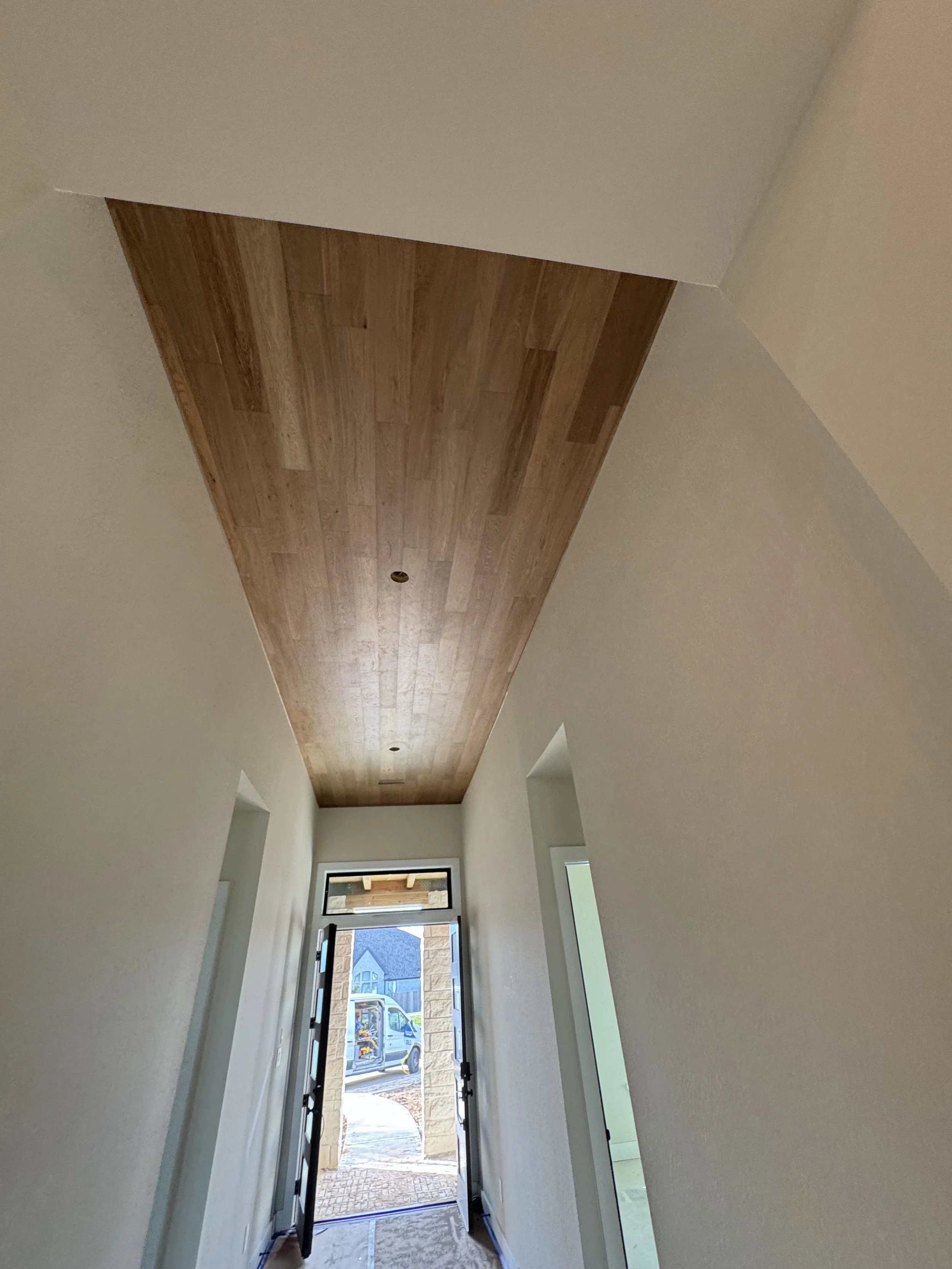 Interior view of a hallway with a wooden ceiling, white walls, and a door at the end leading outside, with construction materials visible.