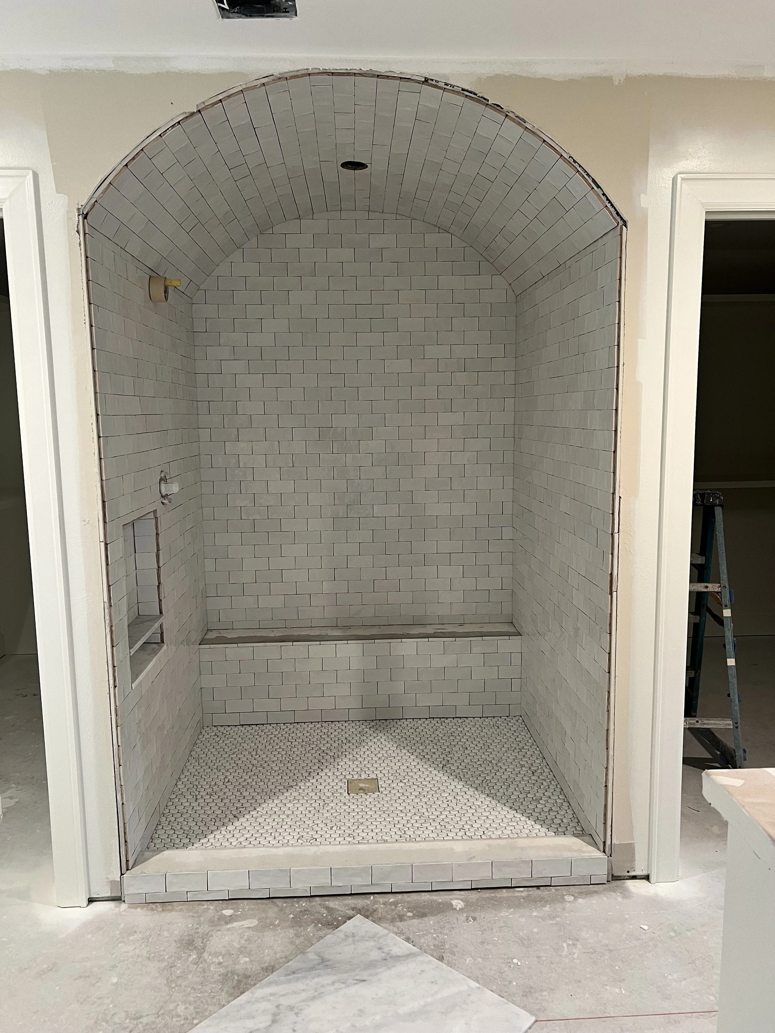 Empty walk-in shower with white subway tile walls, arched top, small hexagon tile floor, and recessed shelf in the wall under construction.