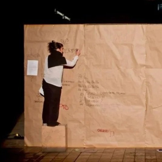 Una mujer escribiendo en una pared de papel marrón en un escenario, con un cubo sobre el que está de pie.