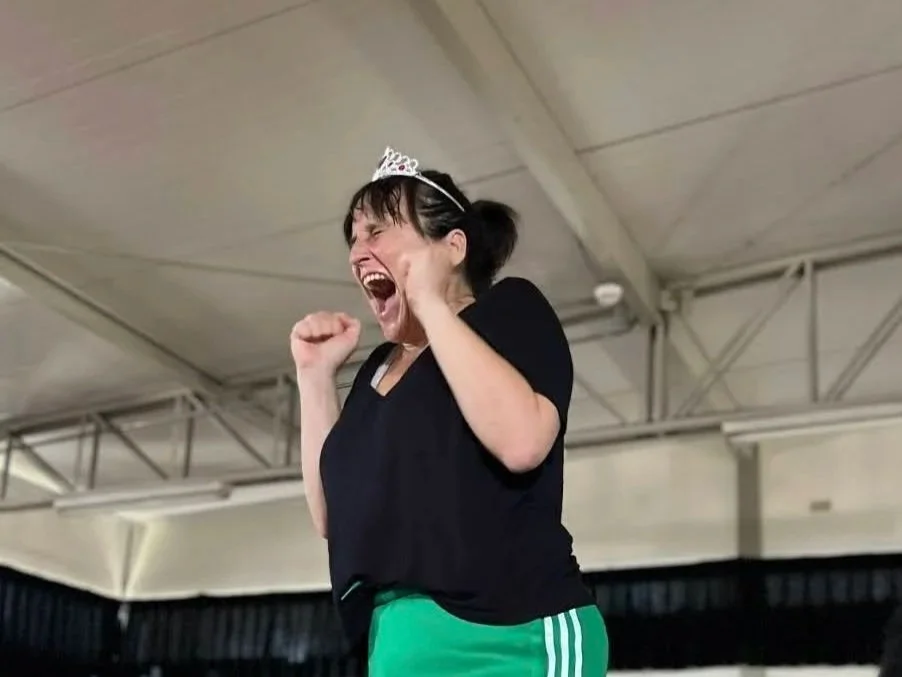 Una mujer con una corona de princesa, vestida con camiseta negra y pantalones deportivos verdes, está gritando felizmente en un espacio cerrado.