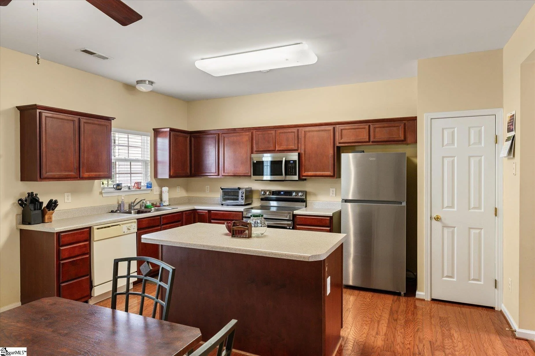 Kitchen with wooden cabinets, a gray refrigerator, a microwave, a stove, a toaster oven, and a window above the sink, with a dining table and chairs in the foreground.