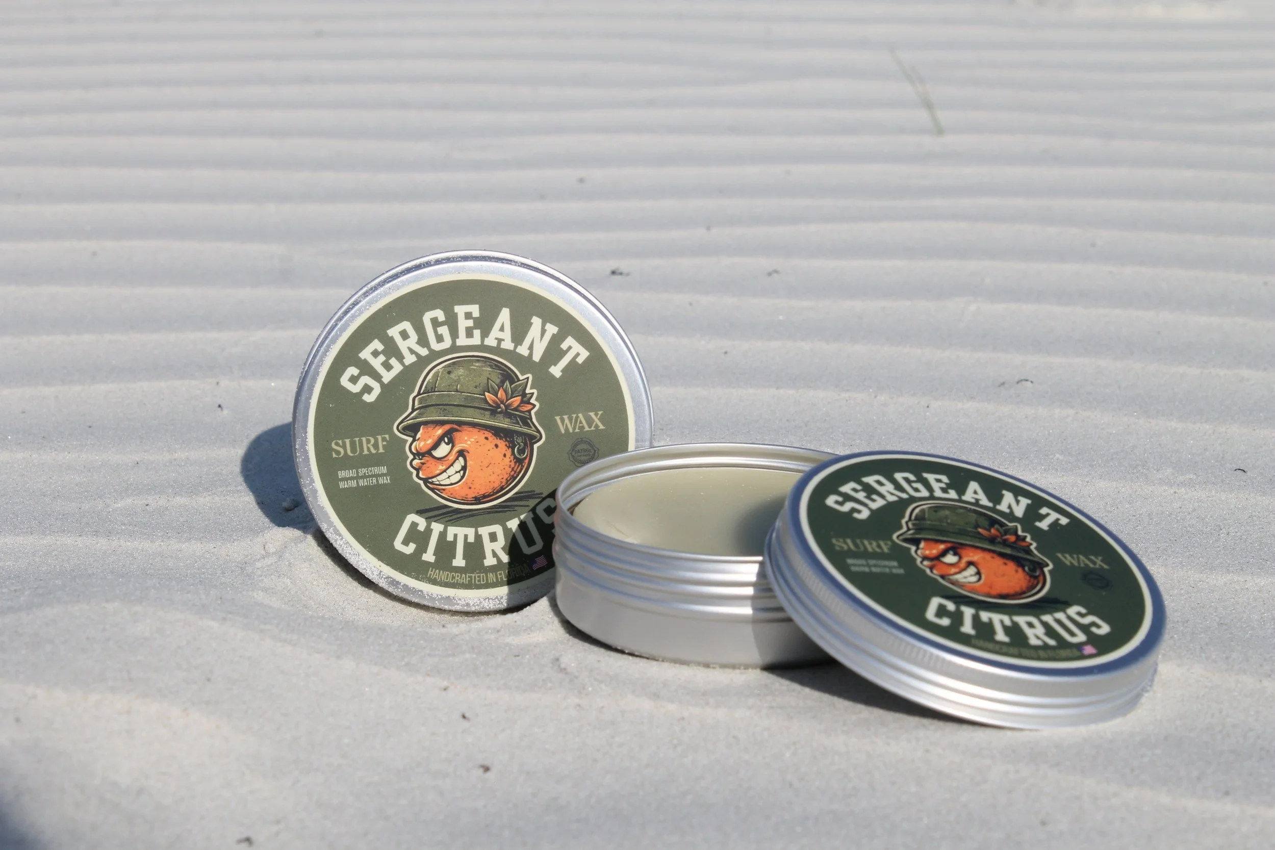 Two tins of Sergeant Citrus surf wax, the left one sitting up in the sand, the right laying flat in the sand with the lid off and surf wax bar exposed.