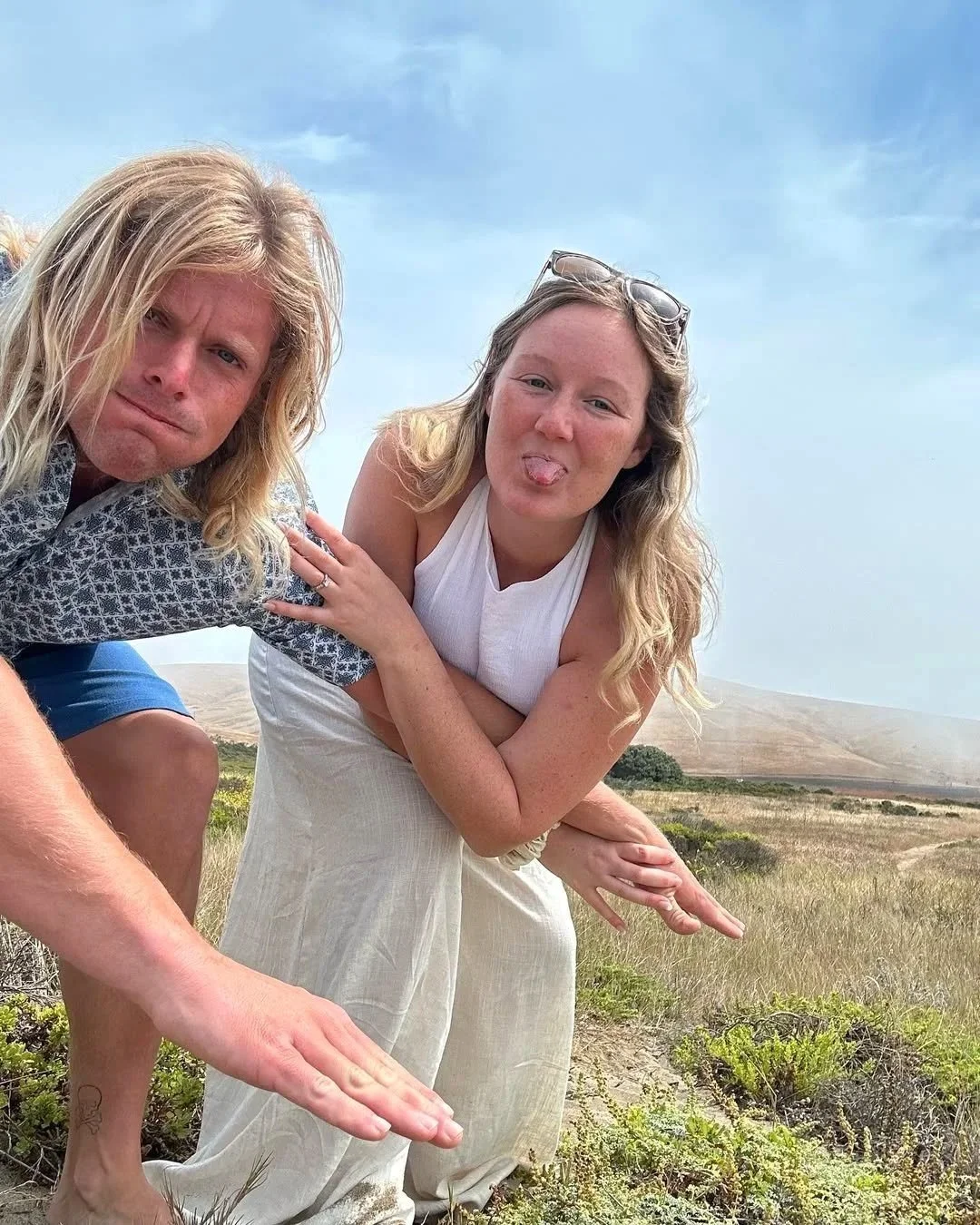 Two women posing outdoors in a grassy field under a cloudy sky, one sticking out her tongue and the other making a serious face, both with long blonde hair.