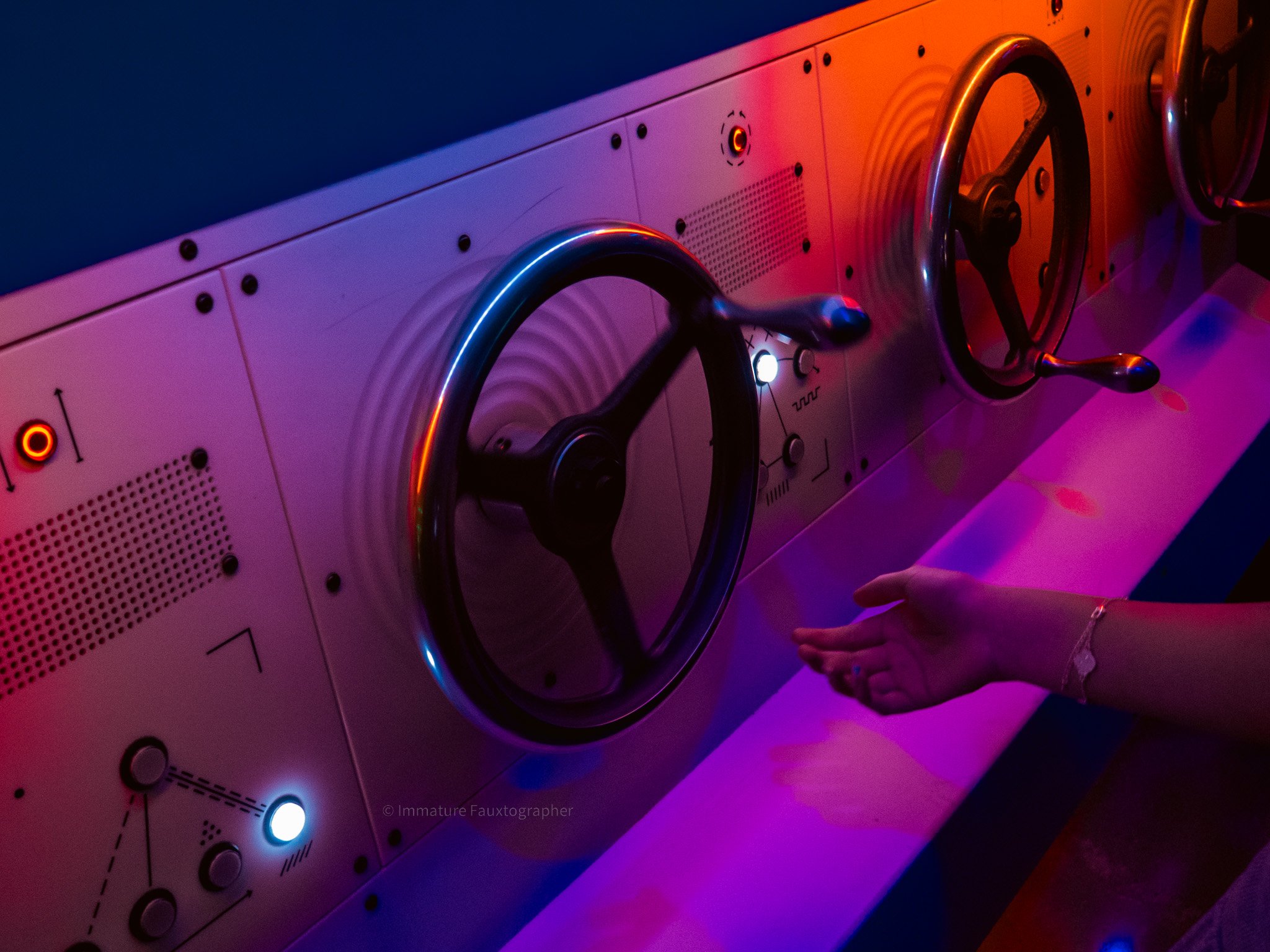 A person's hand reaching towards a control panel with large black knobs under colorful purple and orange lighting.