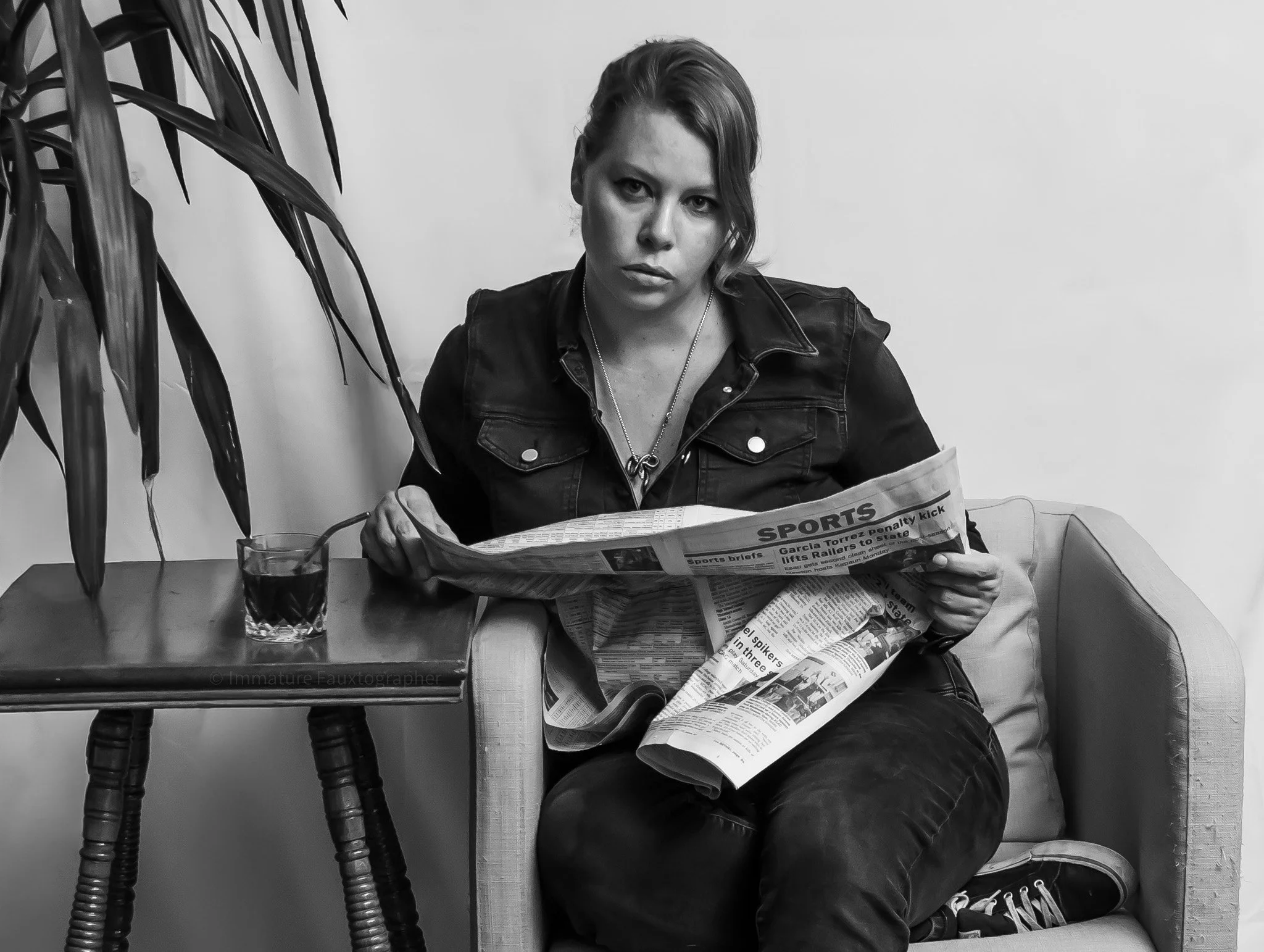 A woman with short hair reading a newspaper, sitting on a couch with a glass of dark beverage and a straw on a small table nearby, in a room with a wall and a plant in the background.