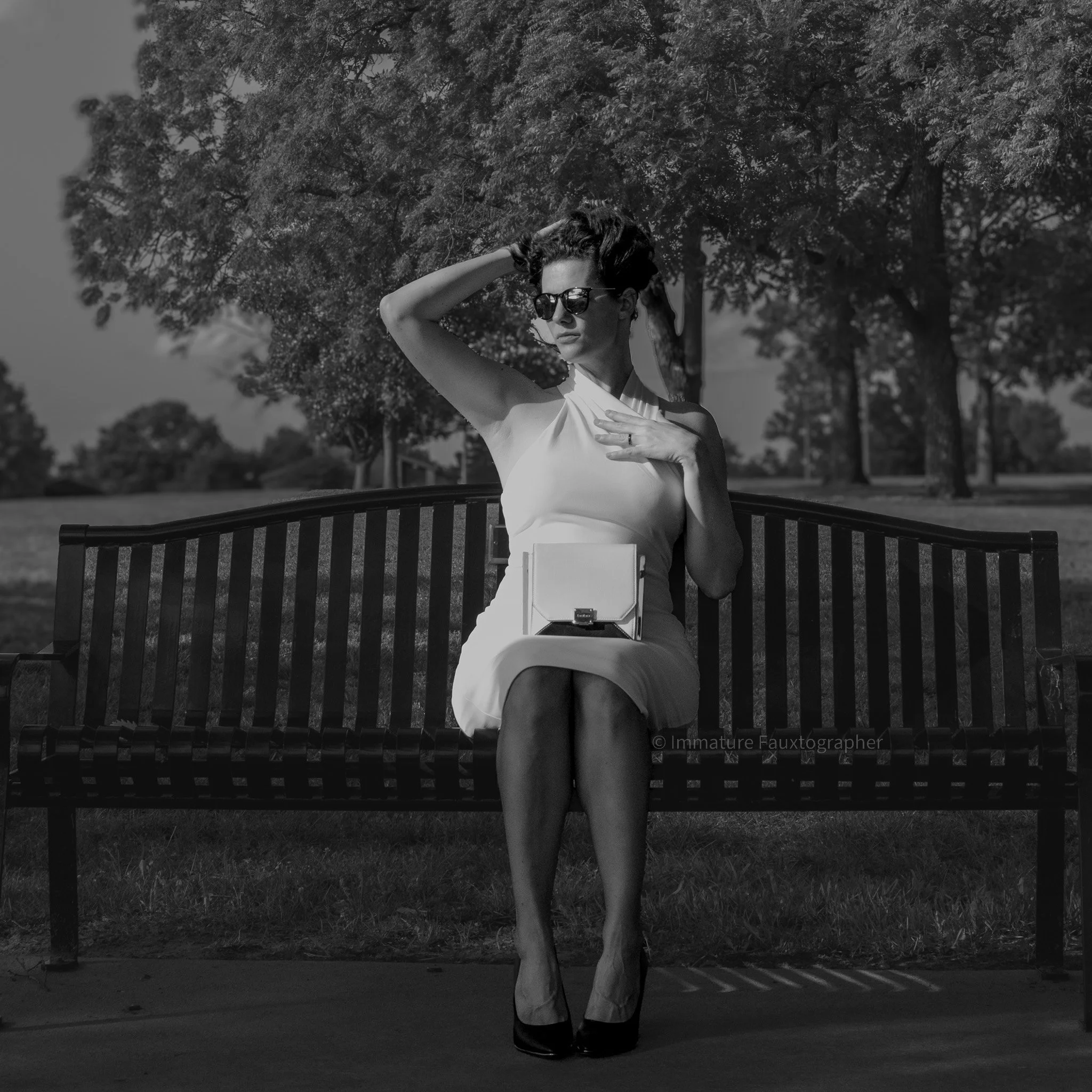 A woman in a white dress sitting on a park bench, wearing sunglasses, with her left hand on her chest and right hand touching her head, in an outdoor park with trees in the background, black and white photo.