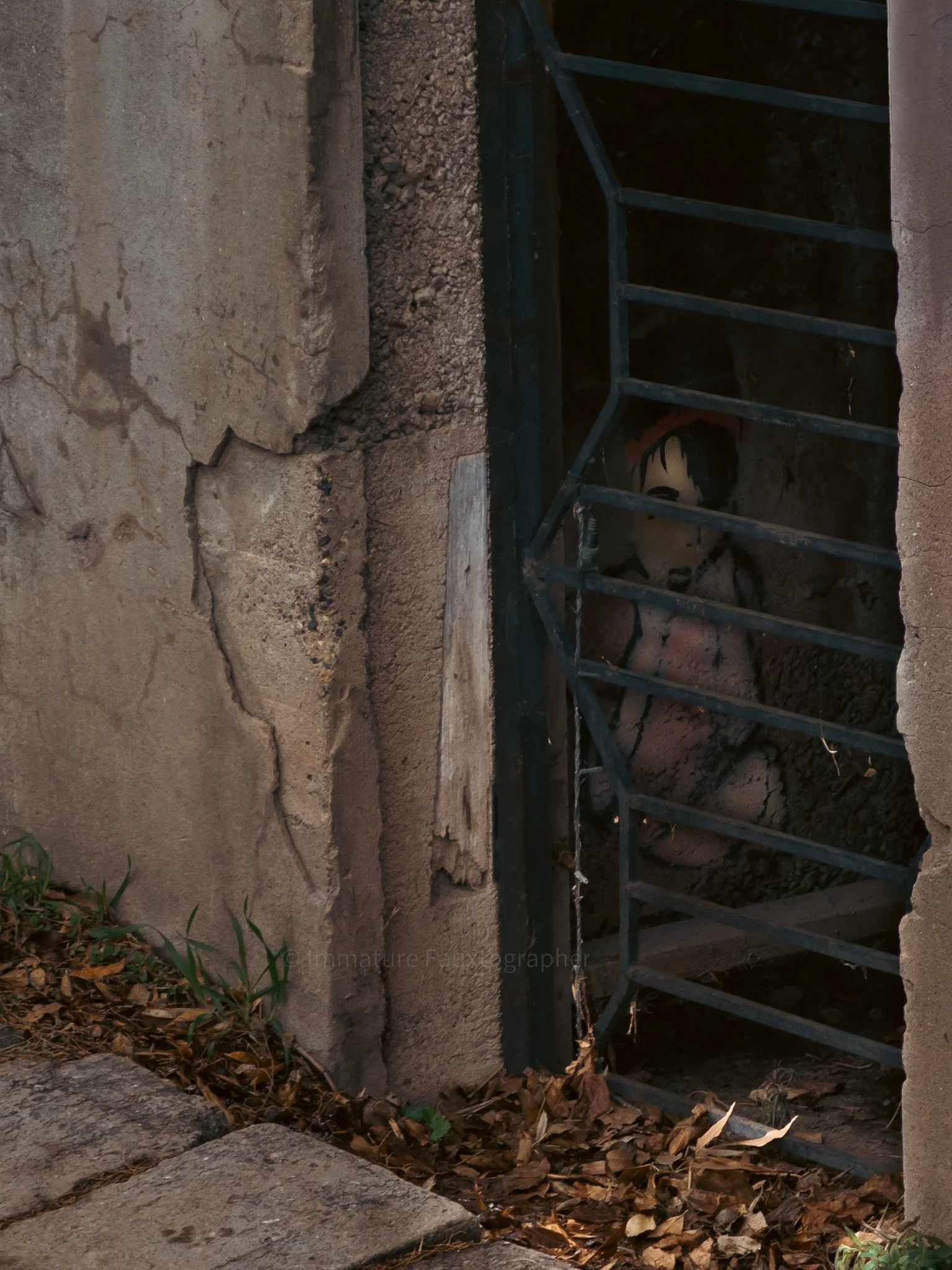 A small, weathered doorway with a child mannequin behind the black metal bars, surrounded by cracked concrete and fallen leaves on the ground.
