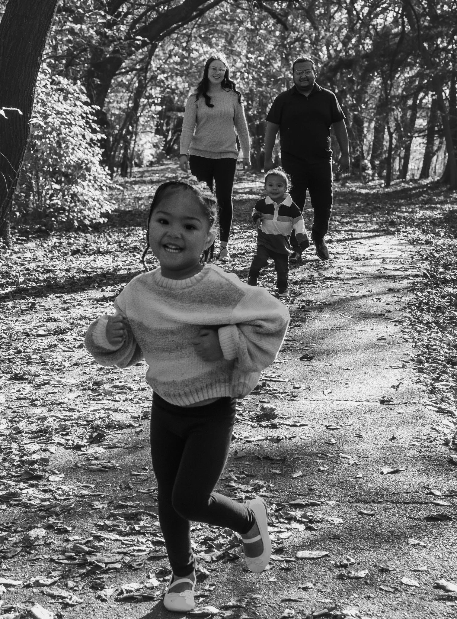 A family of four running on a wooded trail during daytime, with a young girl in the foreground, smiling and wearing a striped sweater and sneakers, and two adults and a smaller child following behind on a sunny day.