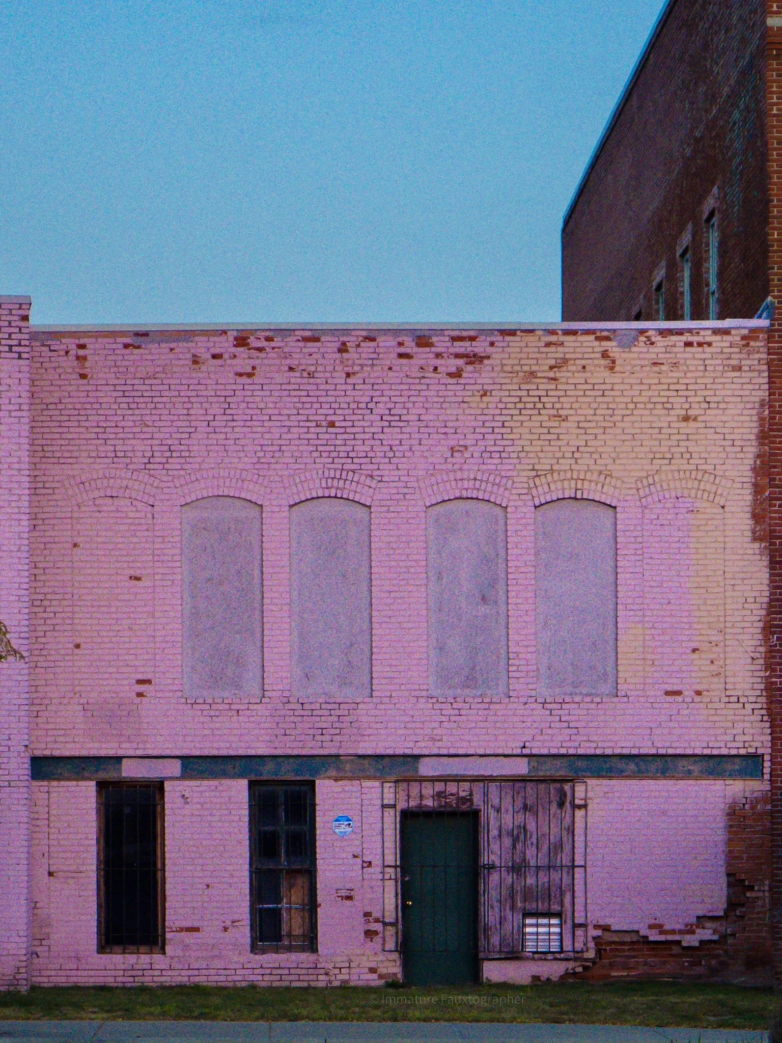 A pink brick building with boarded-up windows and a green door, under a clear blue sky.