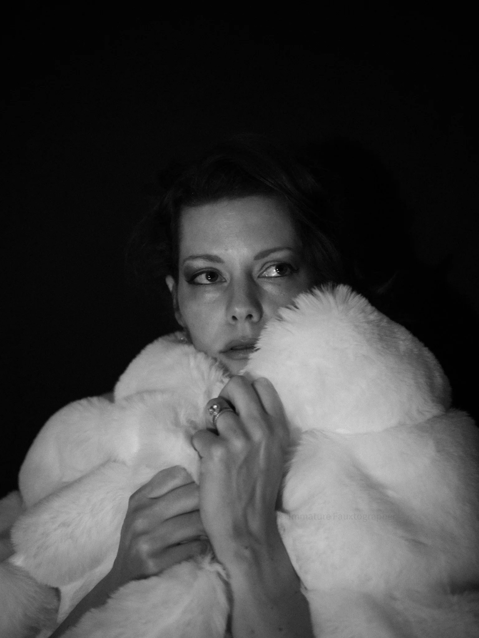 Black and white portrait of a woman wearing a luxurious fur coat, with one hand near her face, gazing to the side against a dark background.