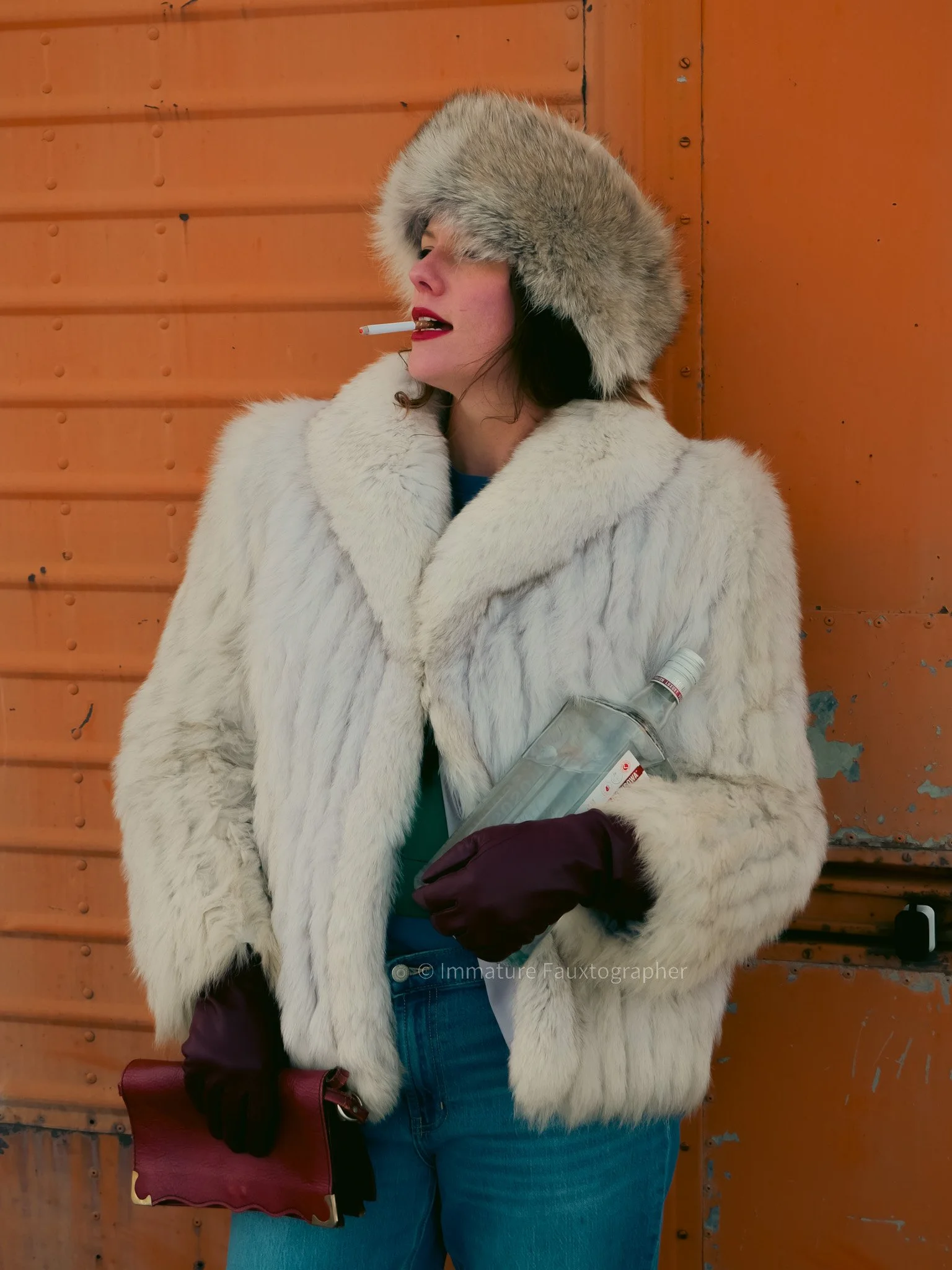A woman wearing a large fur hat and a white fur coat. She has a cigarette in her mouth and is holding a bottle of vodka in her left arm. She is also wearing dark gloves and carrying a burgundy purse, standing against an orange wall, outdoors.