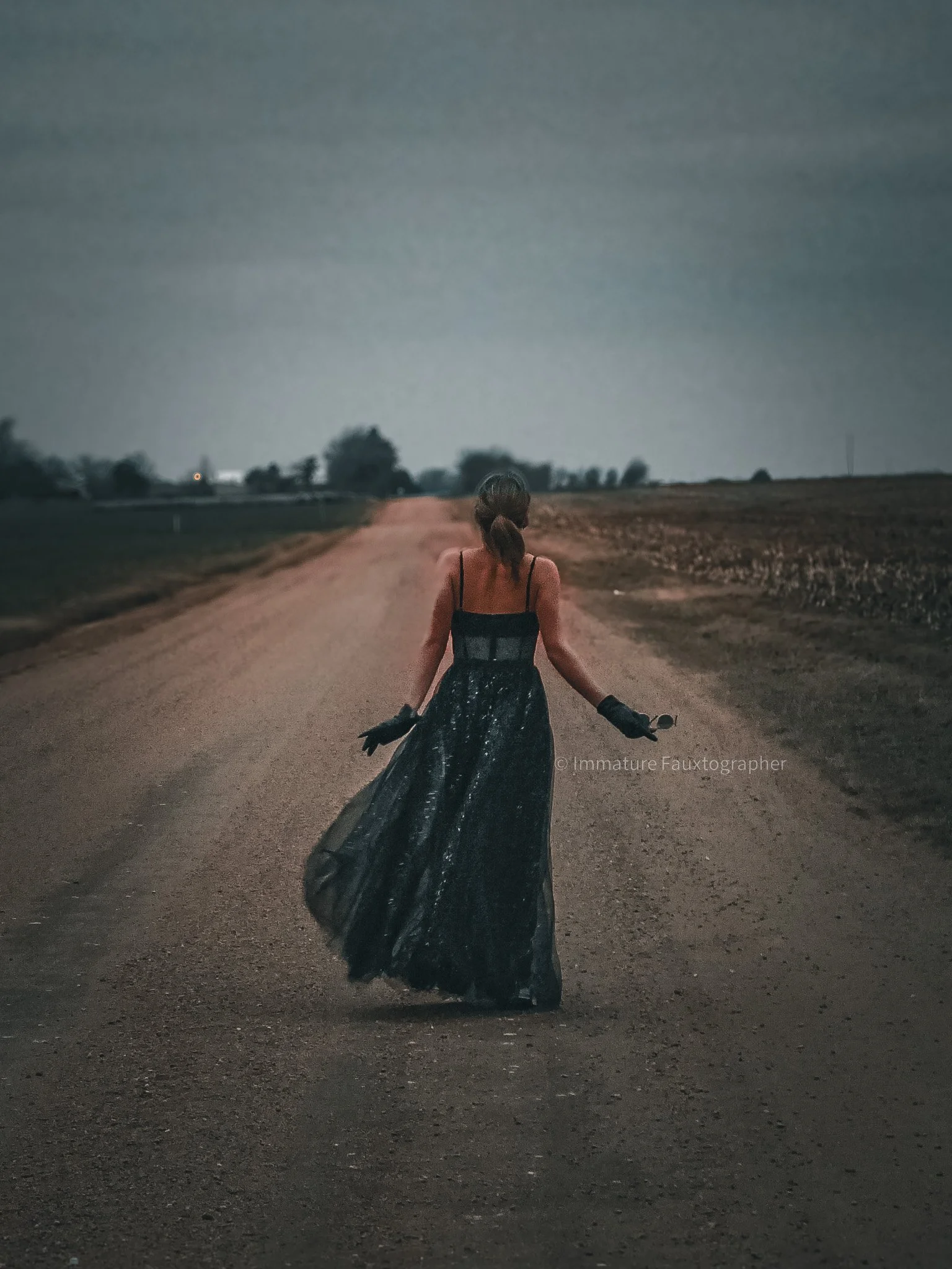 A woman in a black dress walking down a rural dirt road at dusk, with fields on either side and trees in the distance.