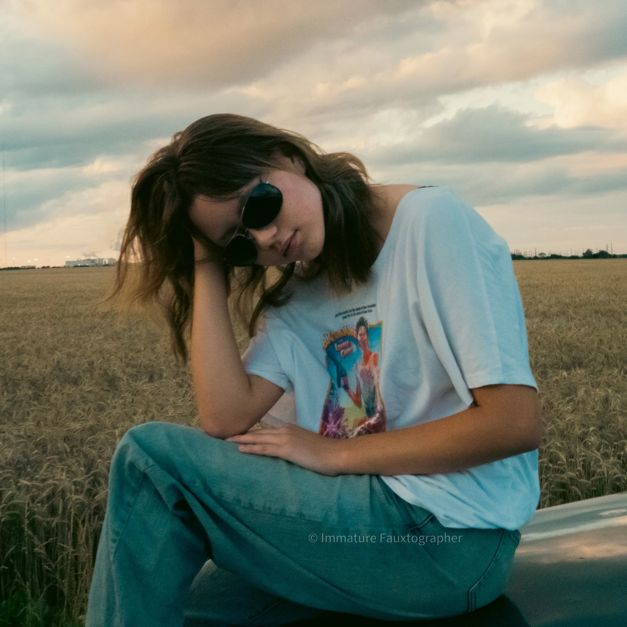 Young woman with shoulder-length brown hair, wearing round black sunglasses, a white graphic t-shirt, and blue jeans, sitting on a car in a rural field during sunset with a cloudy sky in the background.
