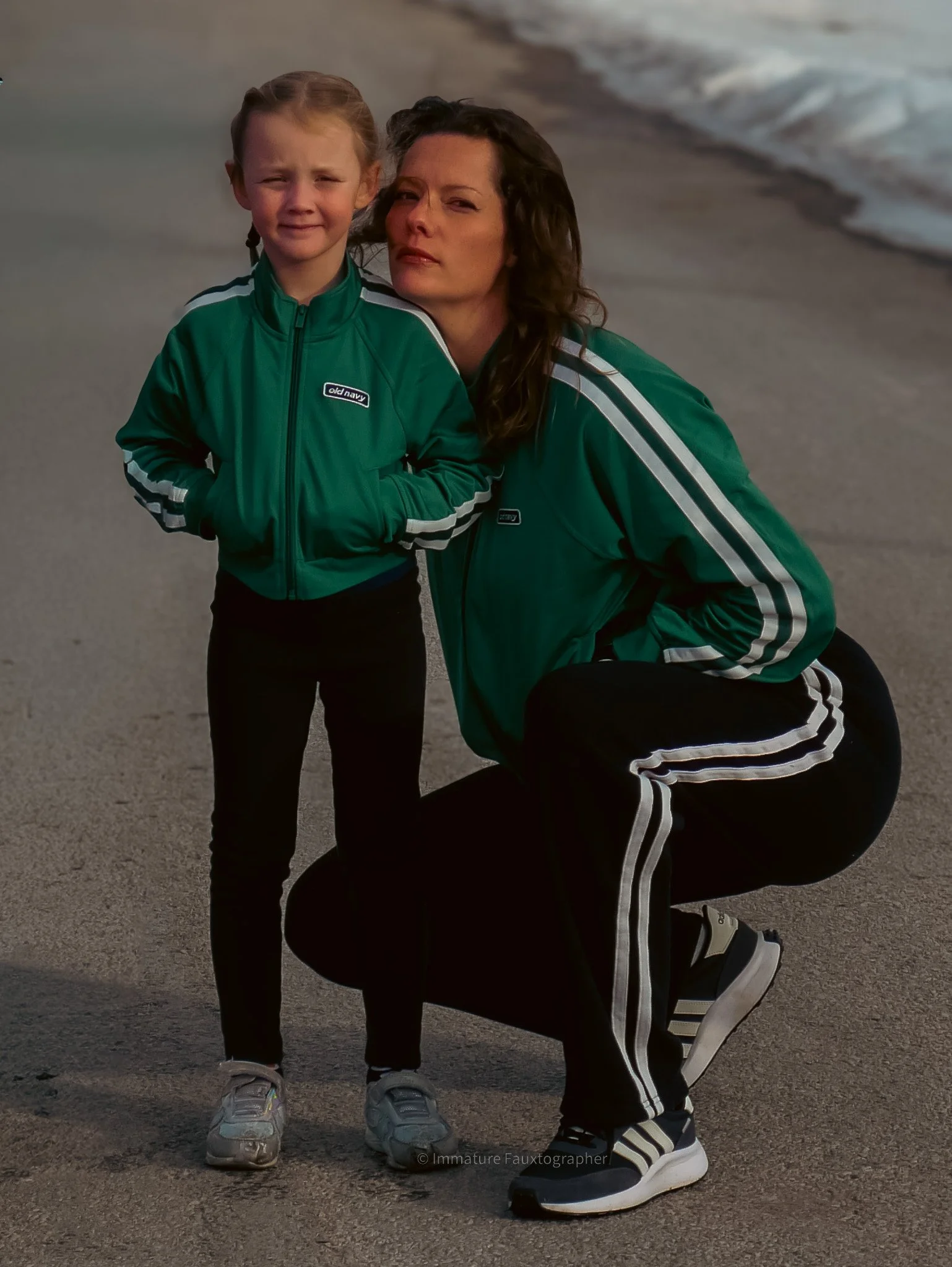 A woman and a young girl on the beach, both wearing green jackets and black pants, with the woman crouching and the girl standing, both looking at the camera with serious expressions.