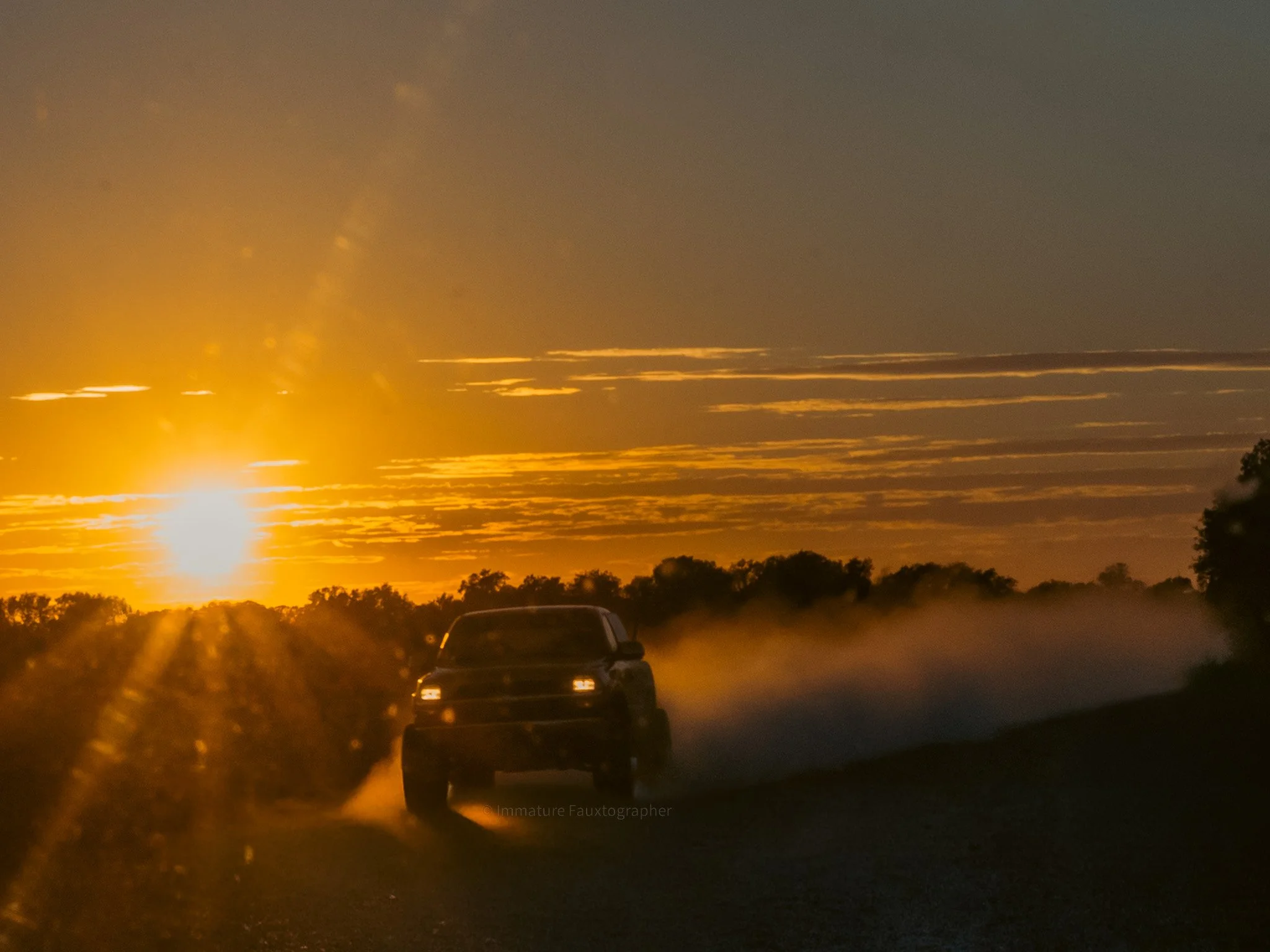 A black pickup truck driving on a dirt road at sunset, with dust clouds behind it and a sky filled with orange and yellow hues.