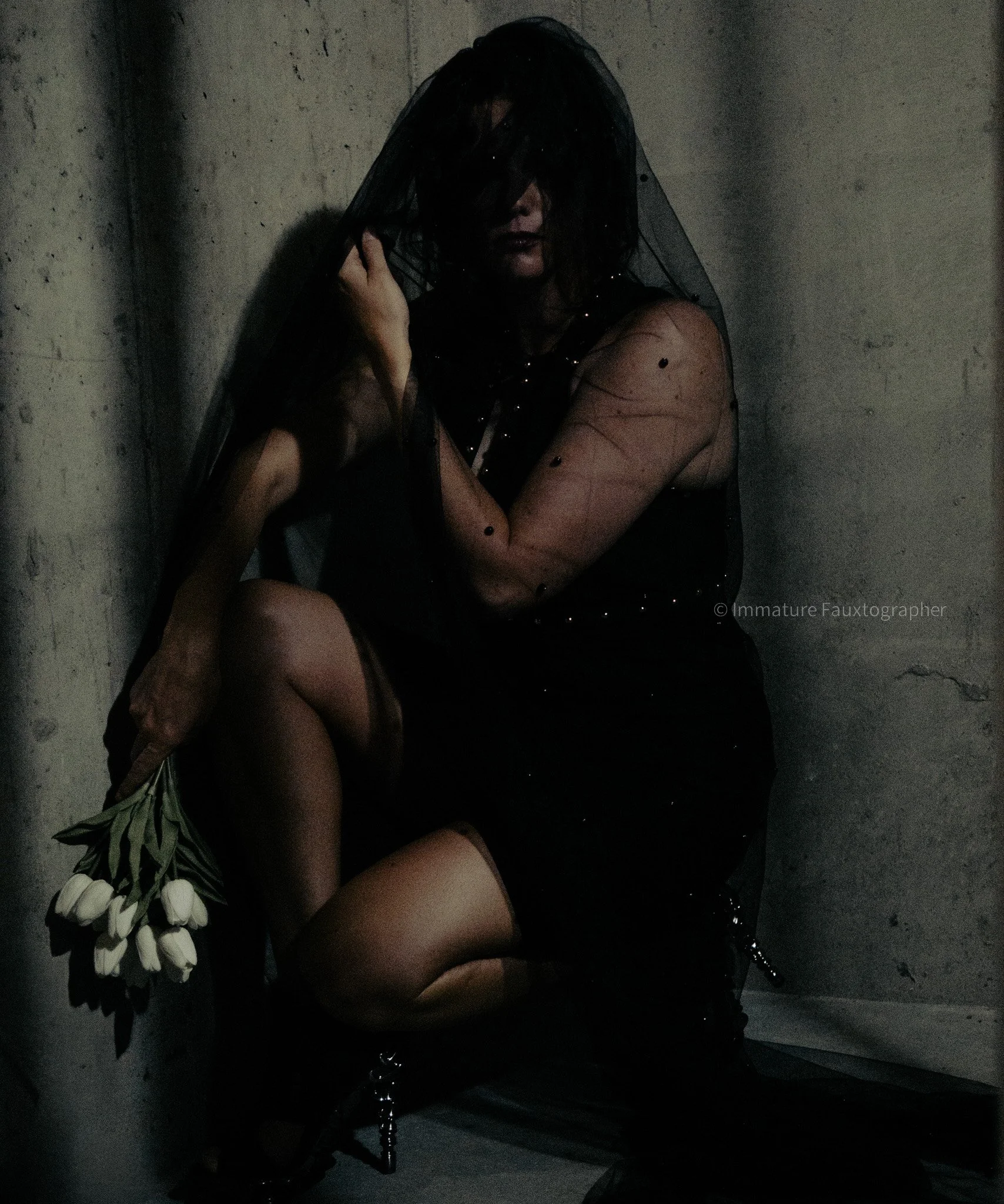 A person dressed in black with a sheer veil covering their face, sitting against a concrete wall, holding a bouquet of white tulips.
