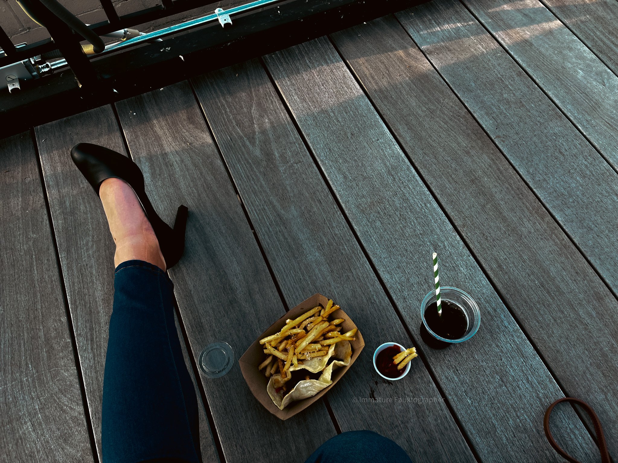 View of a person's leg wearing a black high heel shoe, a container of French fries and a taco, and a drink on a wooden deck.