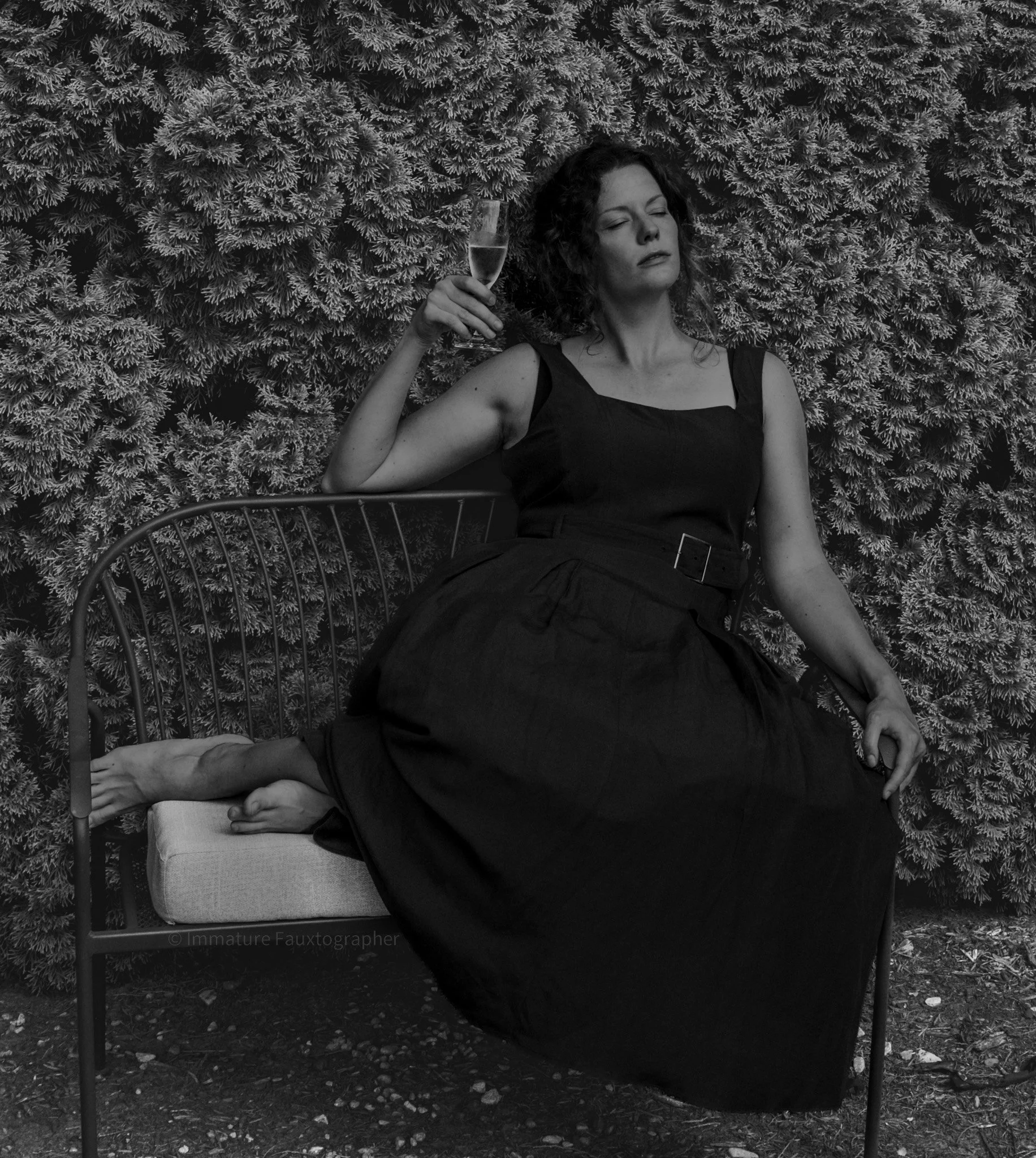 A woman sitting on a metal outdoor bench with a cushion, holding a glass of champagne, with her eyes closed and head tilted back, against a backdrop of dense, leafy shrubs.