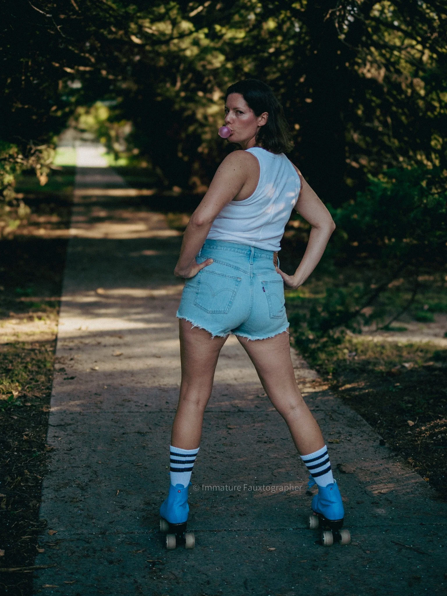 Woman standing on a skateboard on a tree-lined path, blowing a bubble gum bubble, wearing a sleeveless white top, high-waisted denim shorts, striped knee-high socks, and blue roller skates.