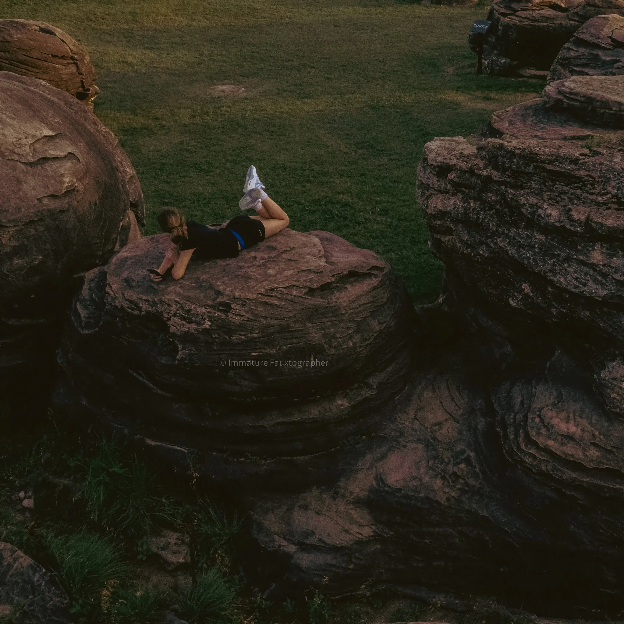 A young woman lying on her stomach on a large, flat rock, looking at her phone, with a grassy area and large rock formations around her in the park.