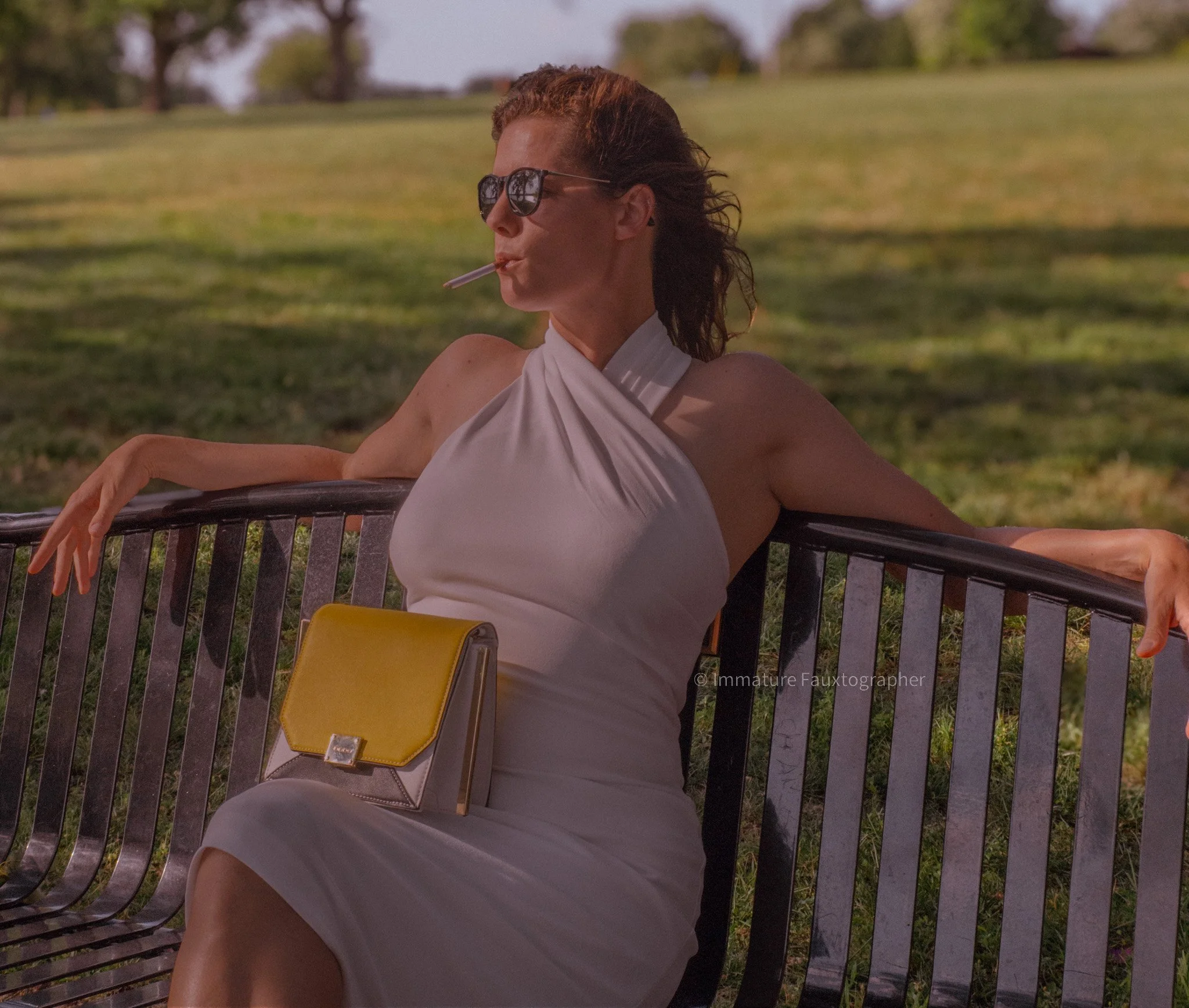 Woman wearing sunglasses, white dress, sitting on park bench, with cigarette in mouth and yellow purse on lap, in a park with trees and grass in the background.