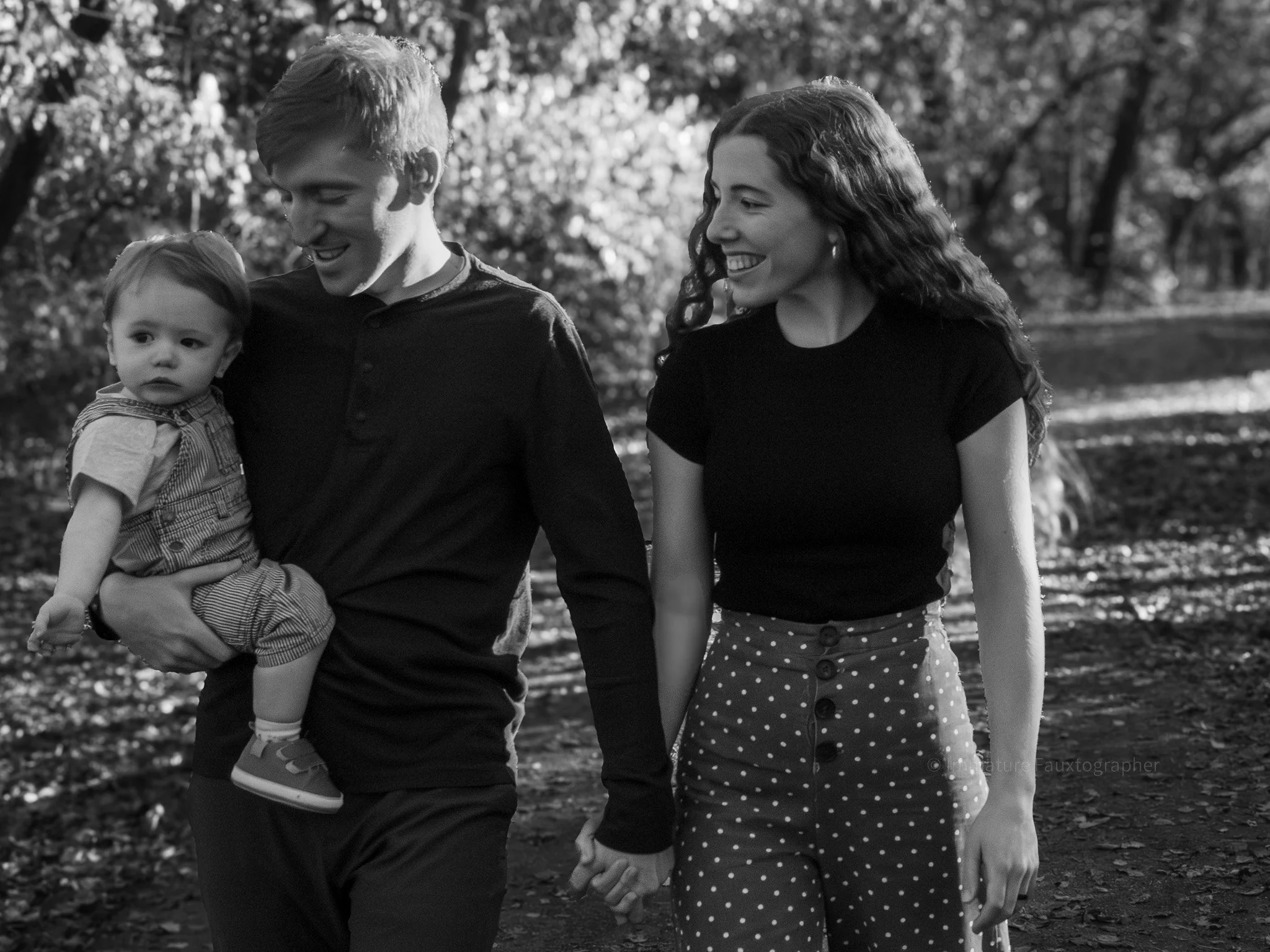 A family of three walking outdoors: a man holding a young child, and a woman holding the man's hand, all smiling and enjoying a sunny day in a park with trees.