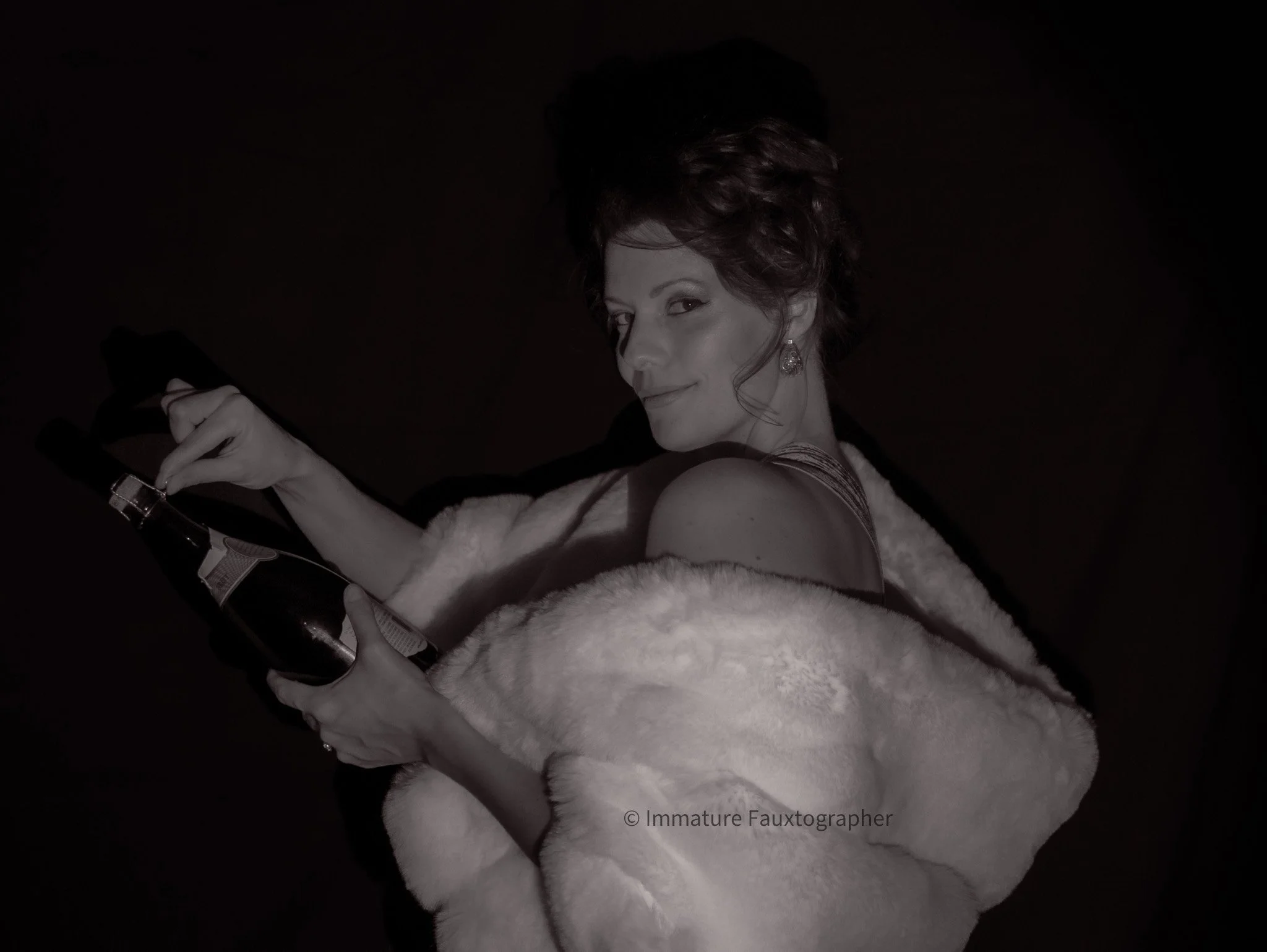 A woman with short, curly hair looks over her shoulder while holding a bottle, wearing a fur coat and a dress with thin straps, against a dark background.