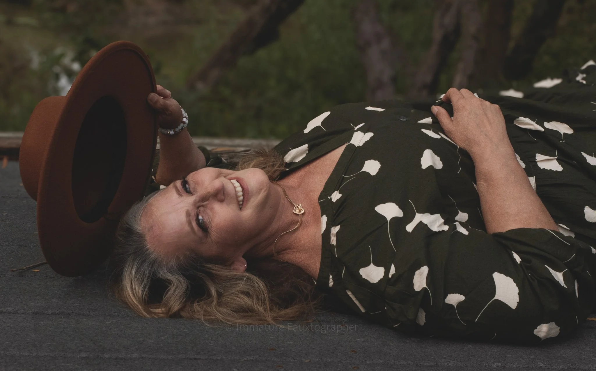 A woman with blonde hair lying on the ground, smiling, holding a brown hat in her right hand, wearing a black dress with white flower prints and a gold heart necklace, outdoors with trees in the background.