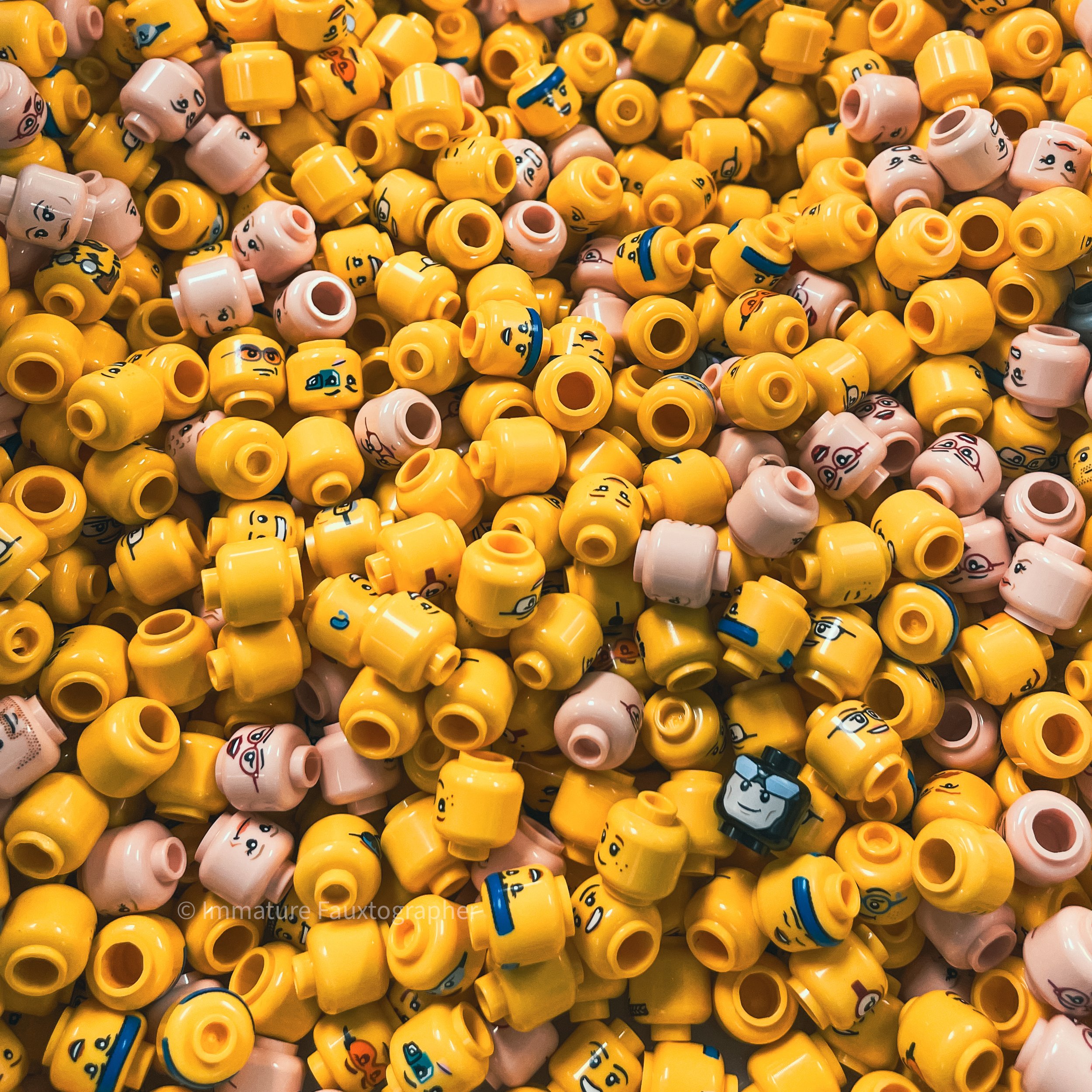 A large pile of small, colorful LEGO minifigure heads, mostly yellow with some pink, featuring various facial expressions and accessories.