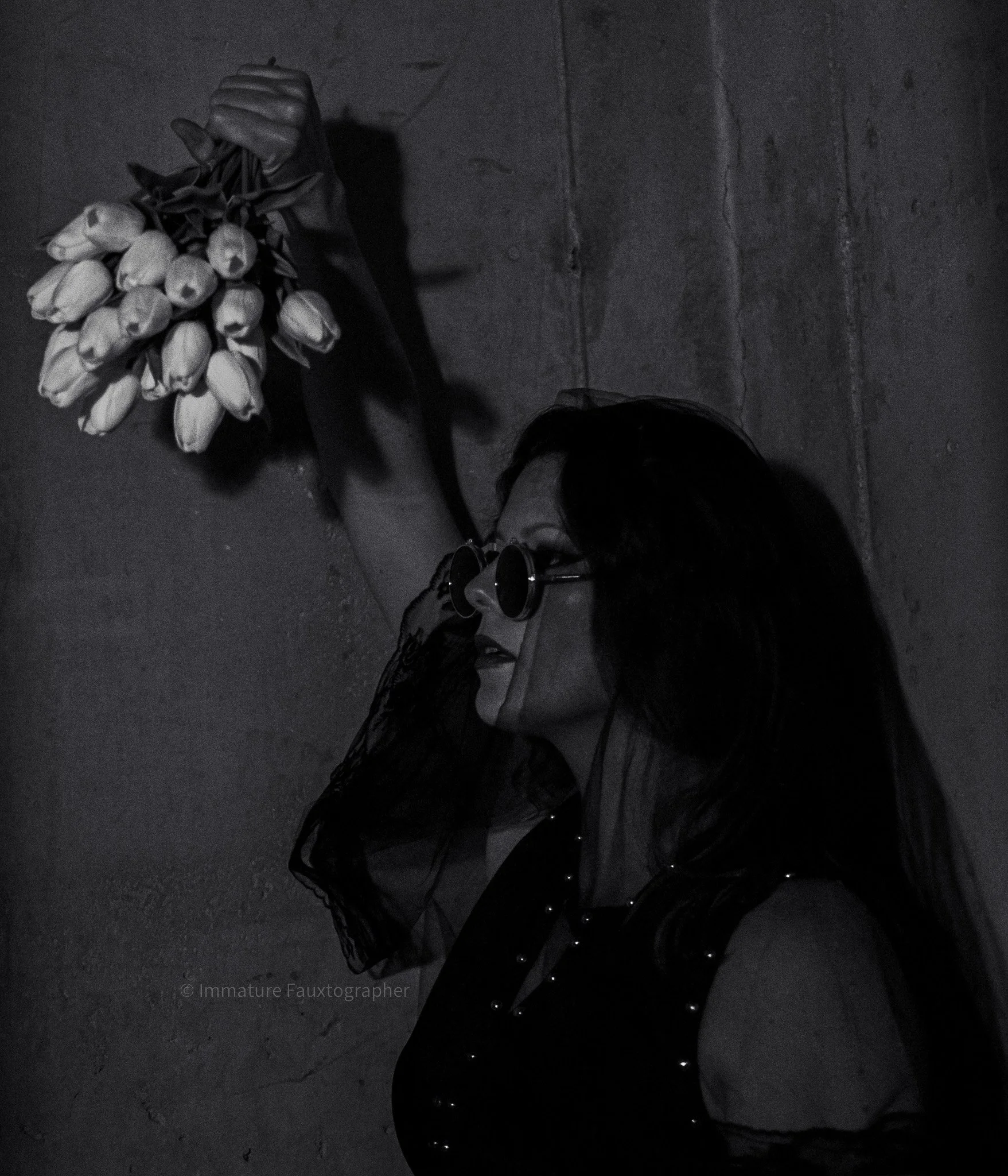 A woman with dark hair, wearing sunglasses and veil, leans against a concrete wall, holding a bouquet of white tulips in her hand.