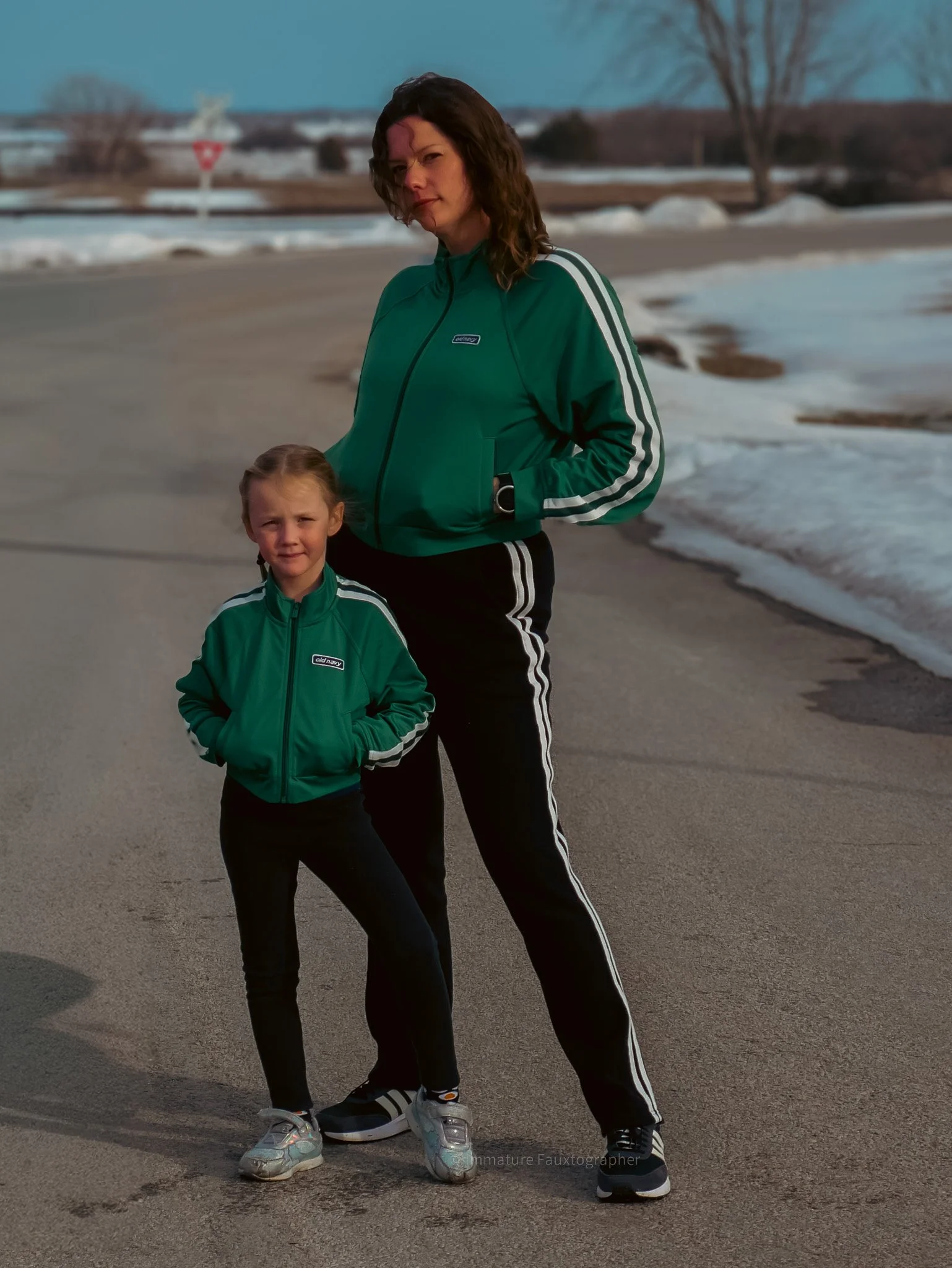 A woman and a girl standing on a paved outdoor area near snow with a road and trees in the background, both dressed in green track jackets and black pants.
