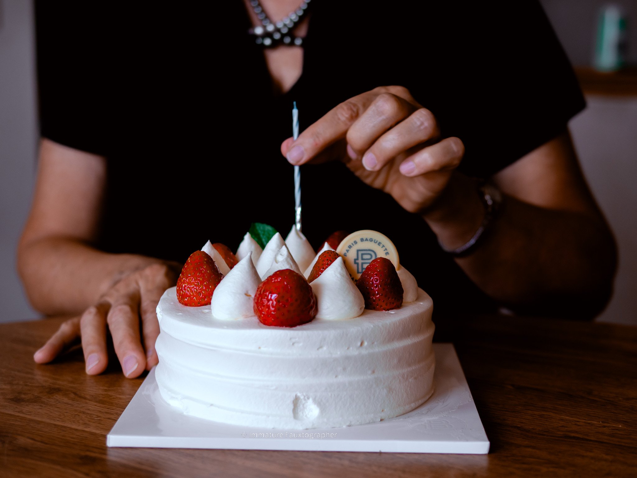 Person in black shirt decorating a white frosted birthday cake with strawberries and a white chocolate disk, using a silver candle lighter.