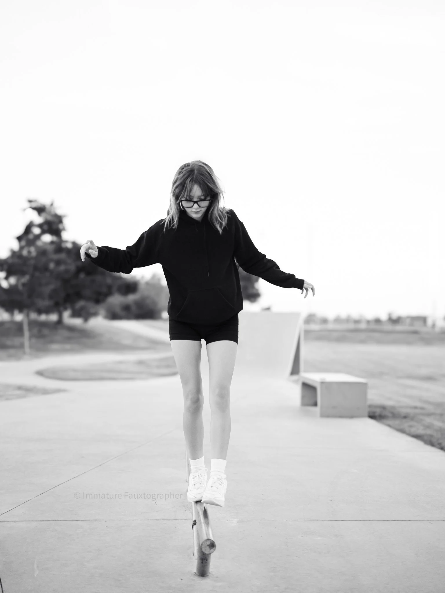 A girl wearing glasses, a black hoodie, and shorts balancing at a skate park.