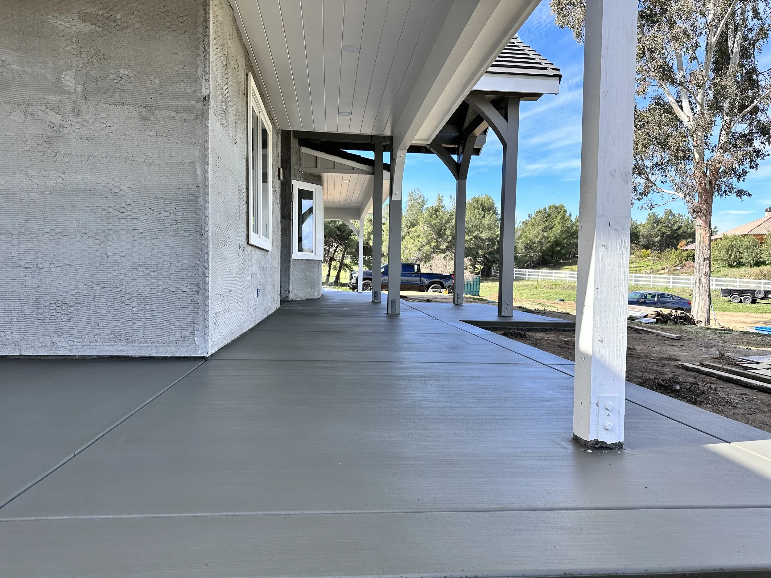 View of a newly constructed house porch with finished concrete floor, white support beams, and a white and gray exterior wall. The background shows trees, a parked truck, and a clear blue sky.