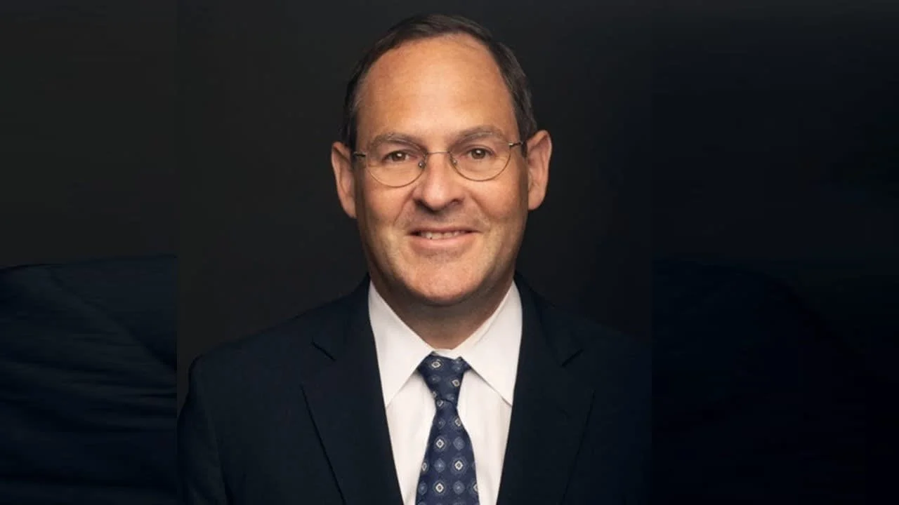 A middle-aged man wearing glasses, a dark suit, white shirt, and patterned tie, smiling at the camera with a dark background.