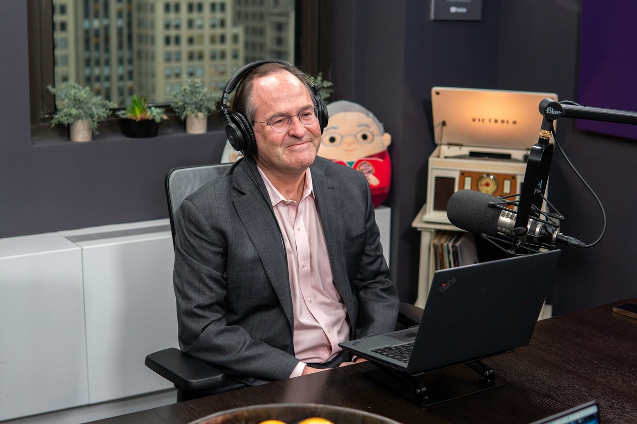 A man in a business suit with glasses and headphones sitting in a recording studio with a microphone, laptop, and decorative items in the background.