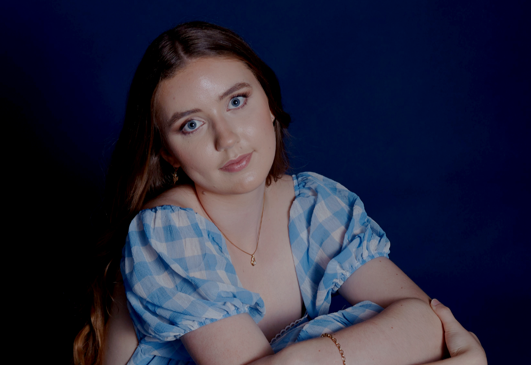 Young woman with blue eyes and long brown hair wearing a blue and white plaid dress, gold necklace with a pearl pendant, and bracelet against a dark background.