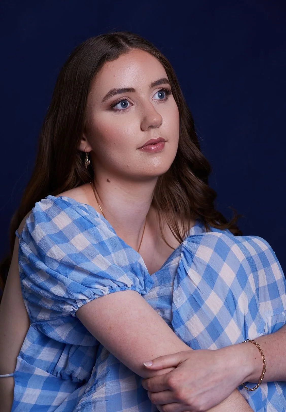 Young woman with long brown hair, blue eyes, wearing a blue and white checkered dress, sitting against a dark blue background.