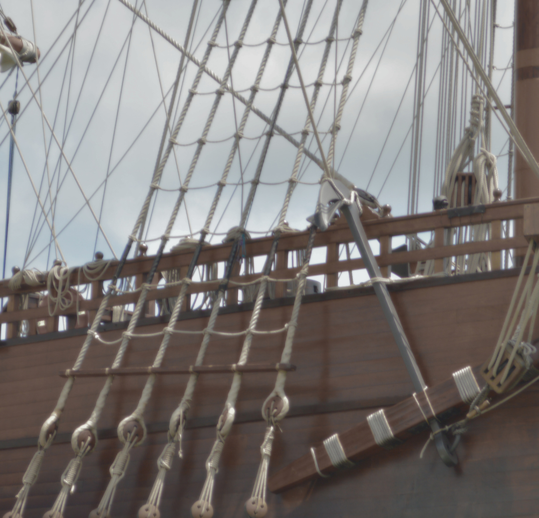 Close-up of a wooden sailing ship's rigging, ropes, and part of the hull against a cloudy sky.