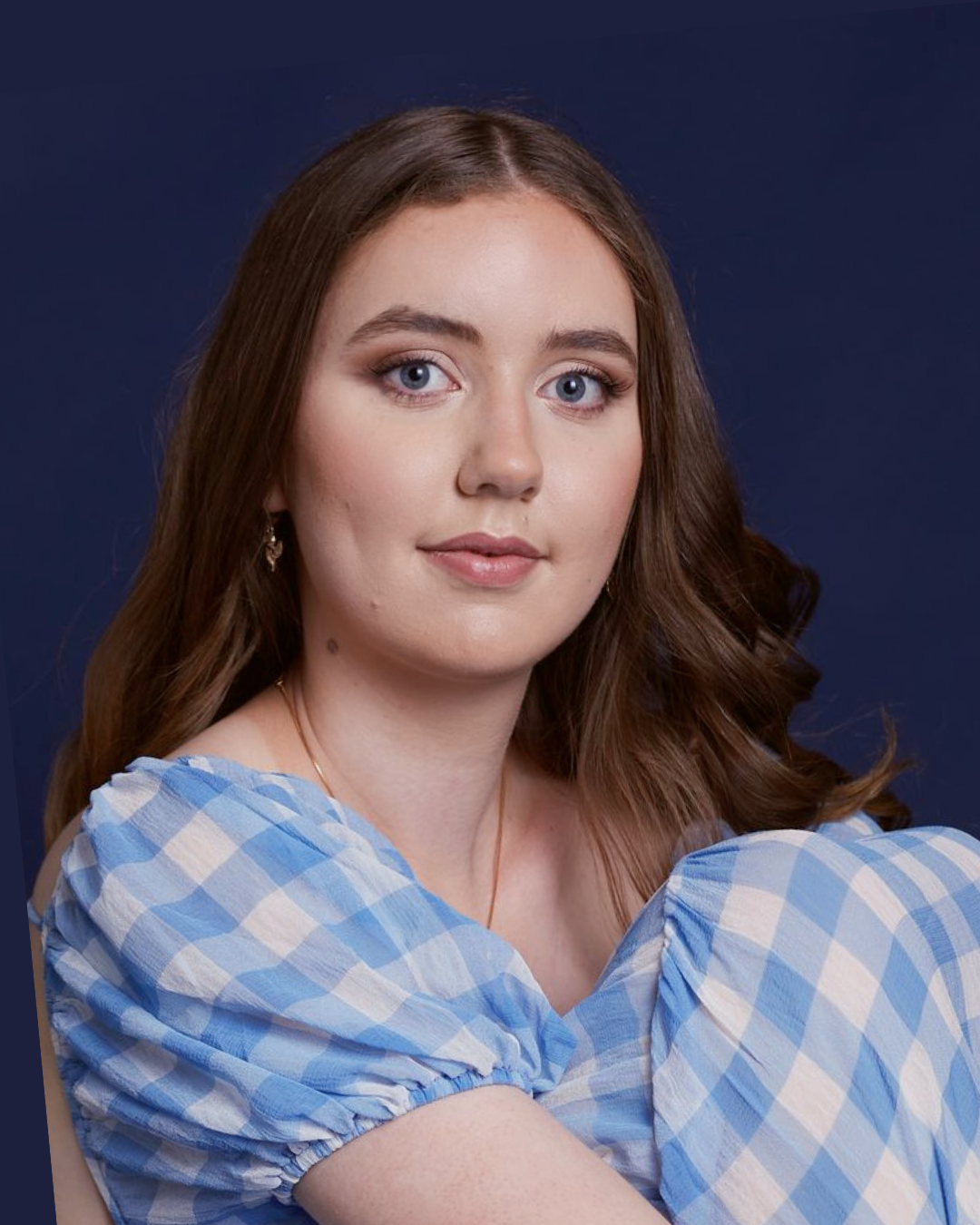 A young woman with long brown hair, blue eyes, and light skin, wearing a blue and white checkered dress, posing against a dark blue background.