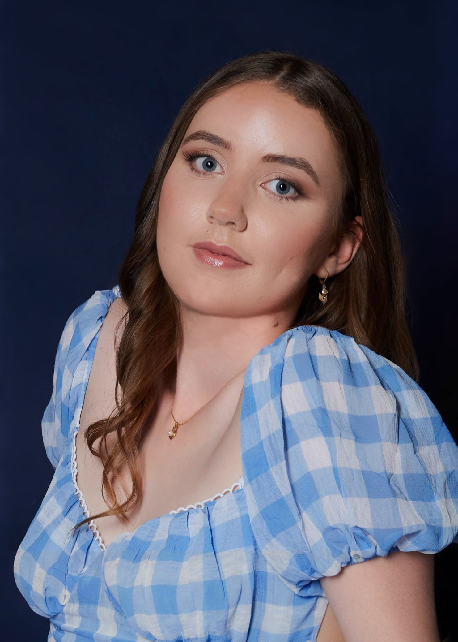 Close-up portrait of a young woman with blue eyes and brown wavy hair, wearing a blue and white checkered off-shoulder top, gold earrings, and a delicate gold necklace with a pearl pendant, against a dark blue background.