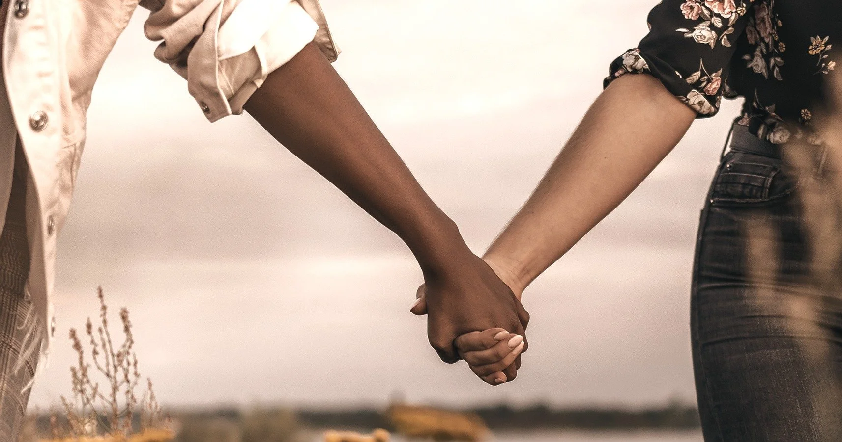 Two people holding hands outdoors with a cloudy sky in the background.